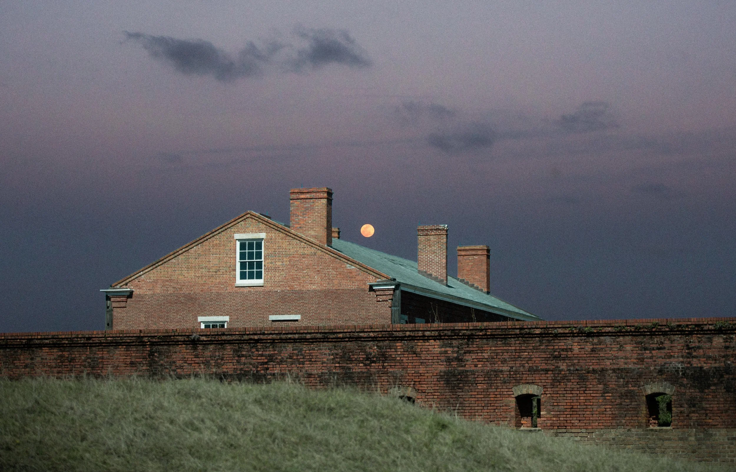A house with a brick exterior and green roof, chimneys, and a window, with the moon visible in the sky and dark clouds above during dusk or dawn.