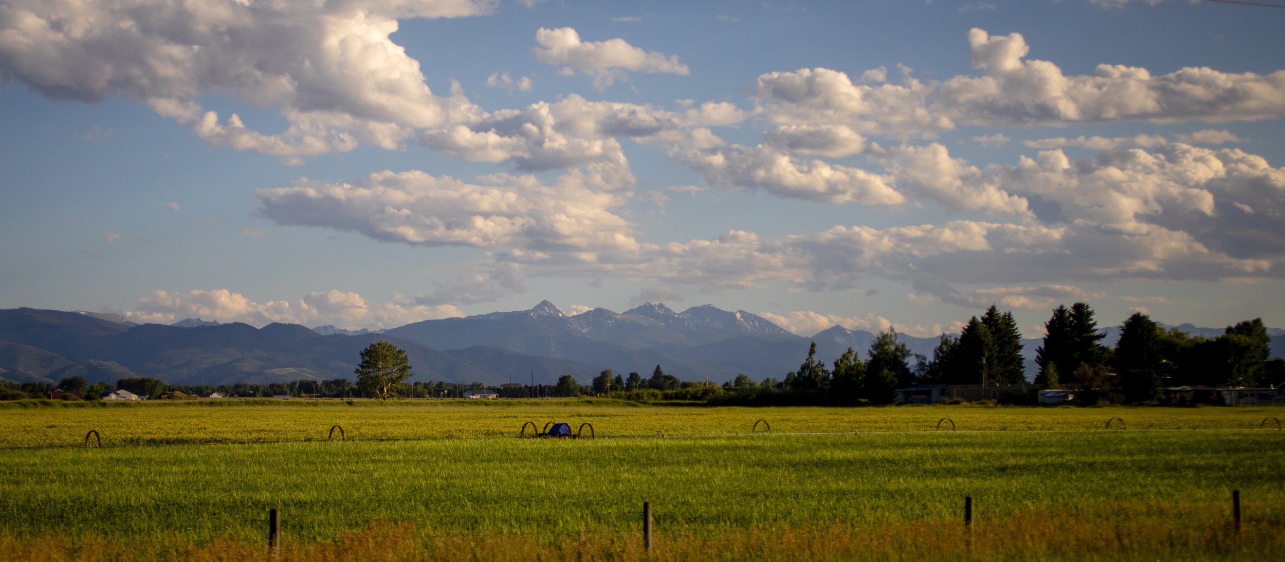 Scenic landscape with green fields, distant mountains with snow-capped peaks, and a partly cloudy sky.
