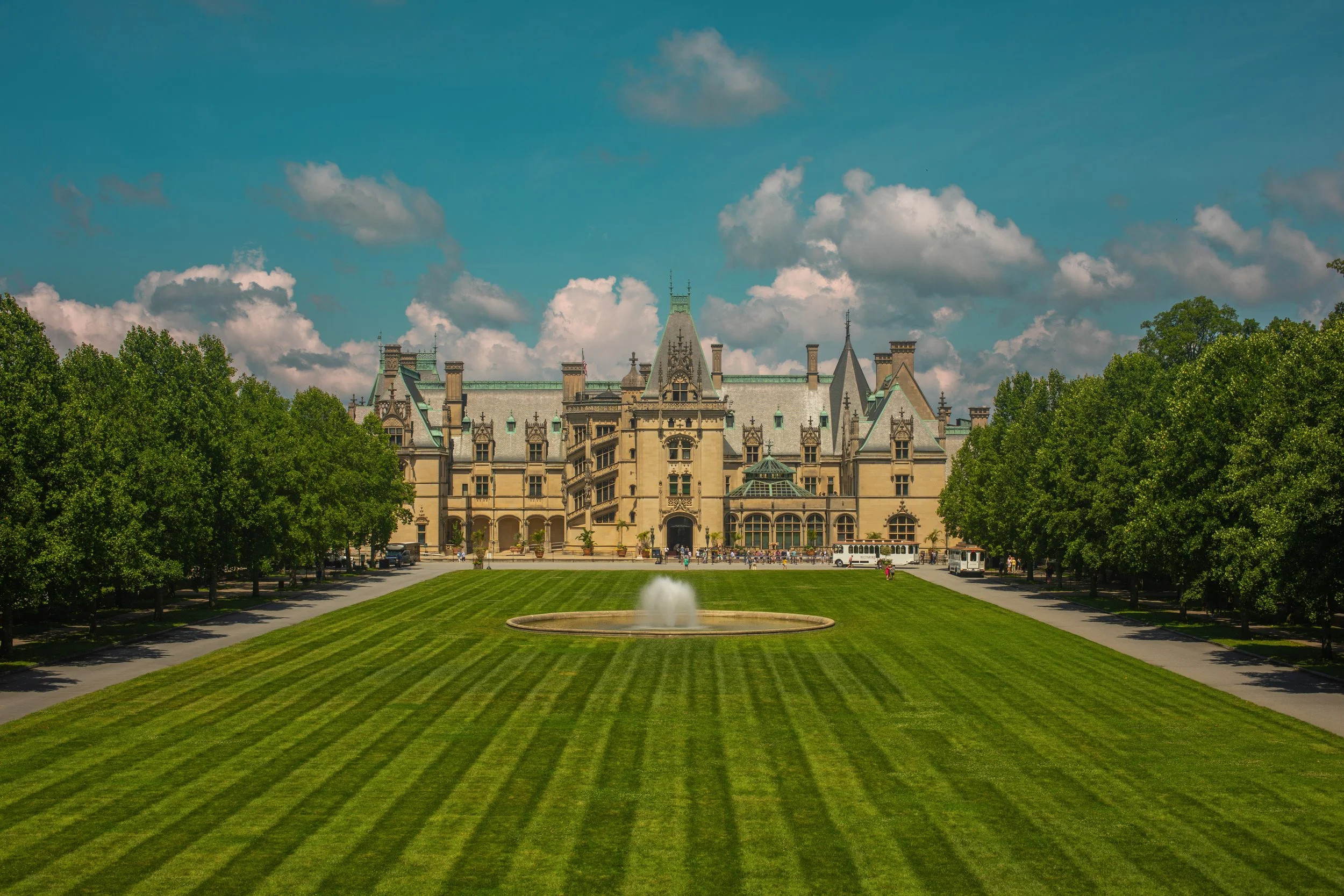A grand historic building with Victorian architecture featuring green rooftops, surrounded by lush green trees and a well-maintained lawn with a fountain in the foreground, under a bright blue sky with scattered clouds.