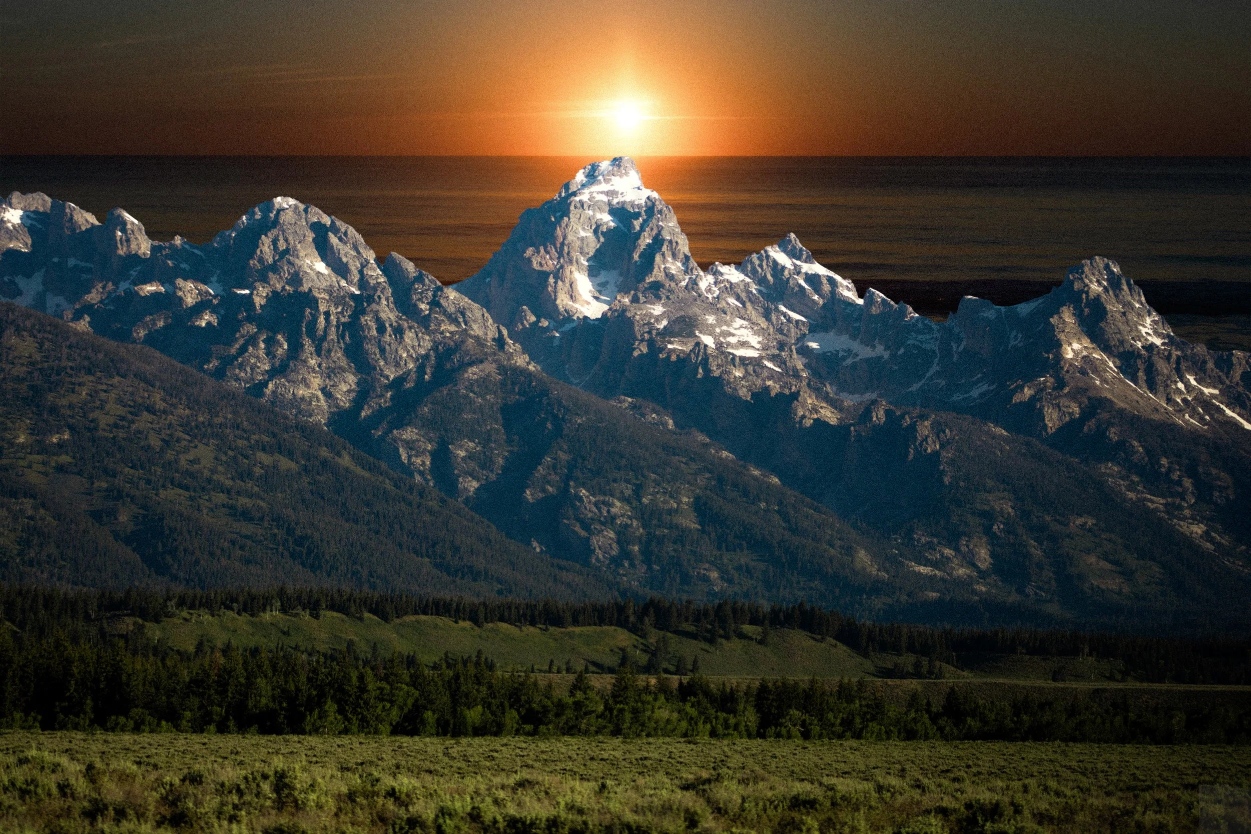 Snow-capped mountain range with a sun setting behind the peaks, green fields in the foreground.