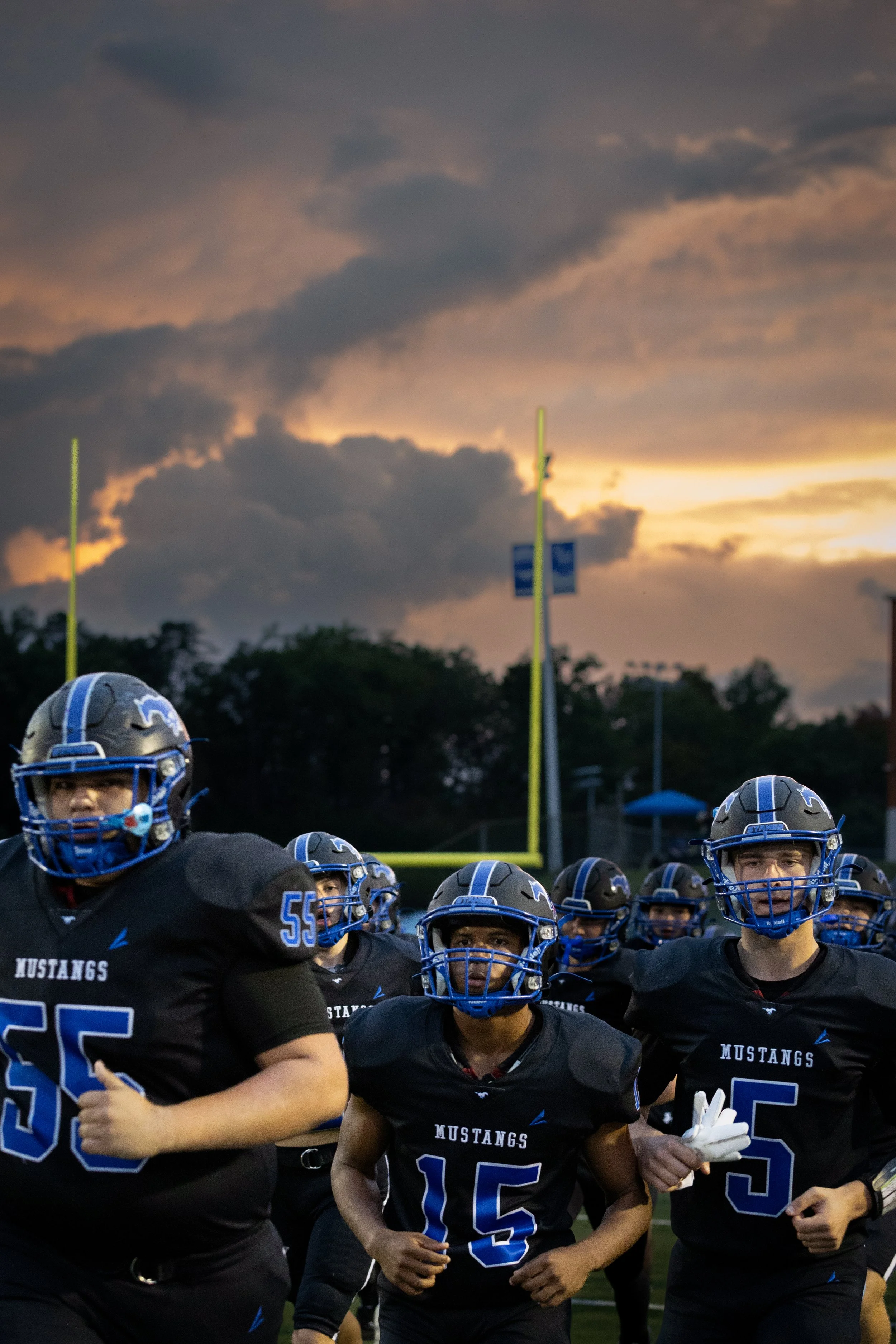 Group of football players, wearing black jerseys with 'Mustangs' and blue numbering, standing on a football field during sunset, with cloudy sky and goalposts in the background.