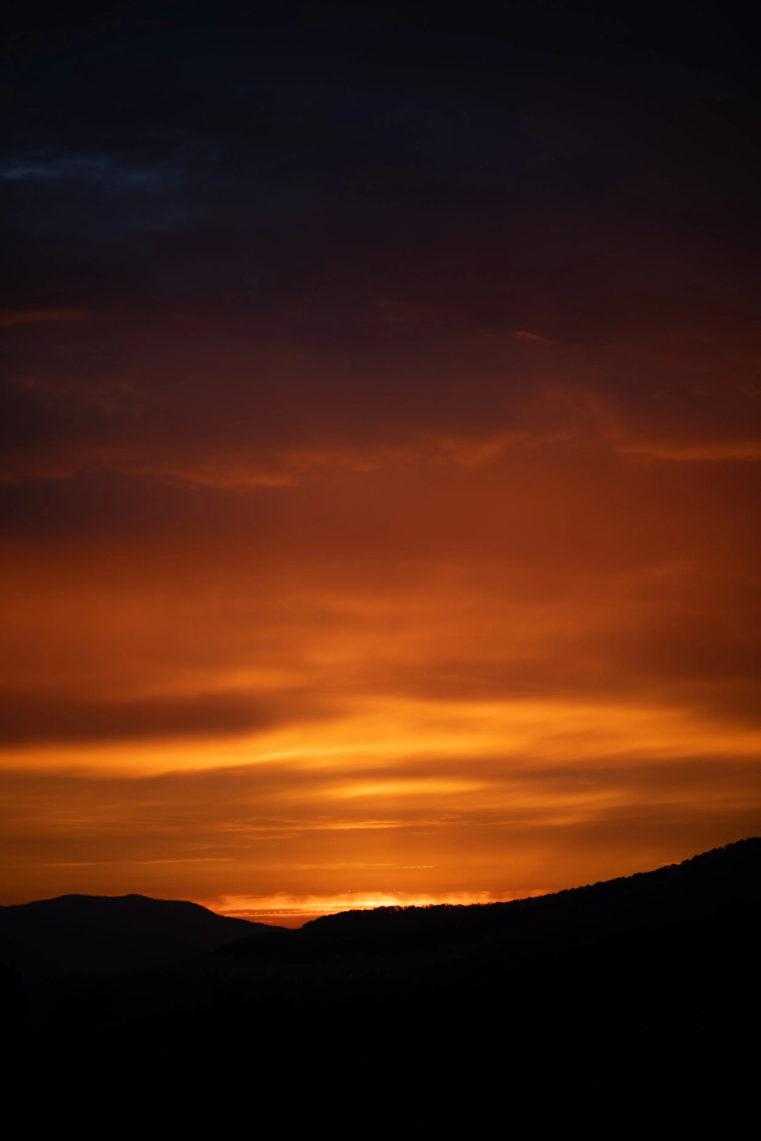 Sunset over mountains with dark silhouetted terrain and colorful sky with shades of orange, yellow, and dark blue.