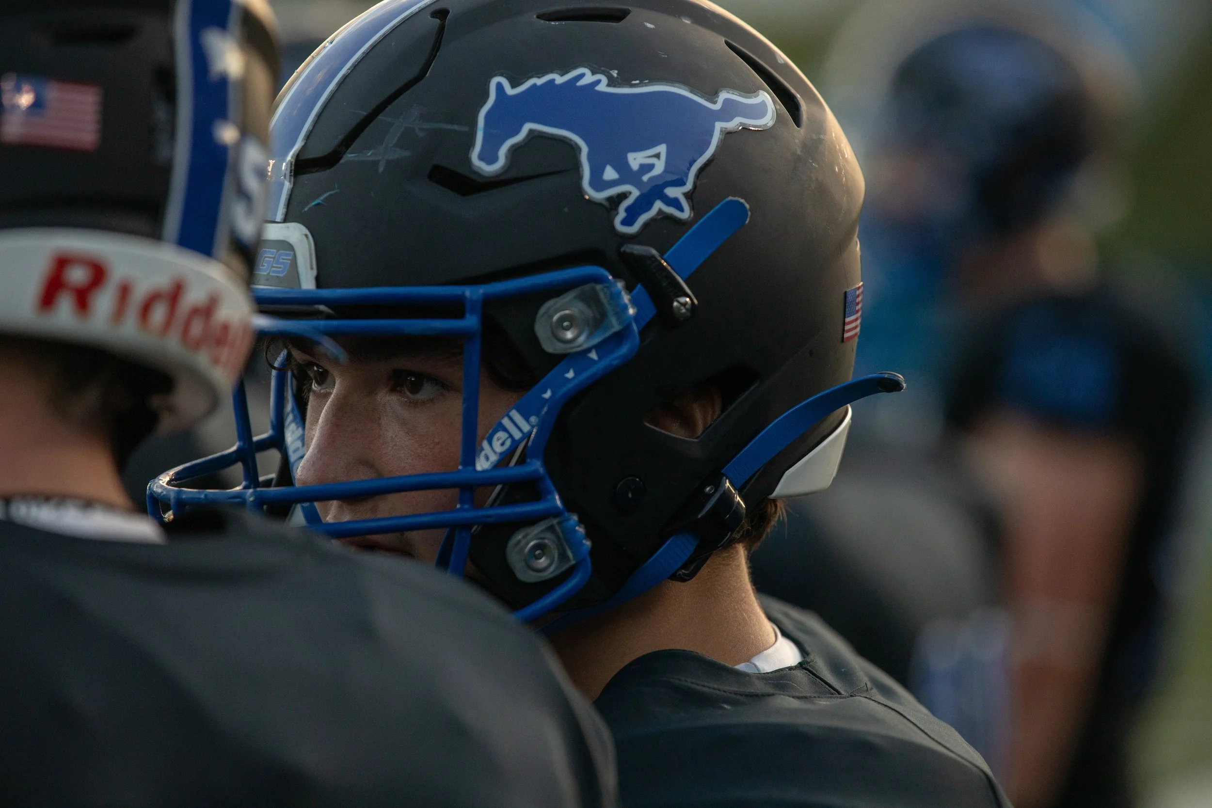 Close-up of a person wearing a black football helmet with a Detroit Lions logo, looking at another player wearing a black helmet with a Raiders logo.
