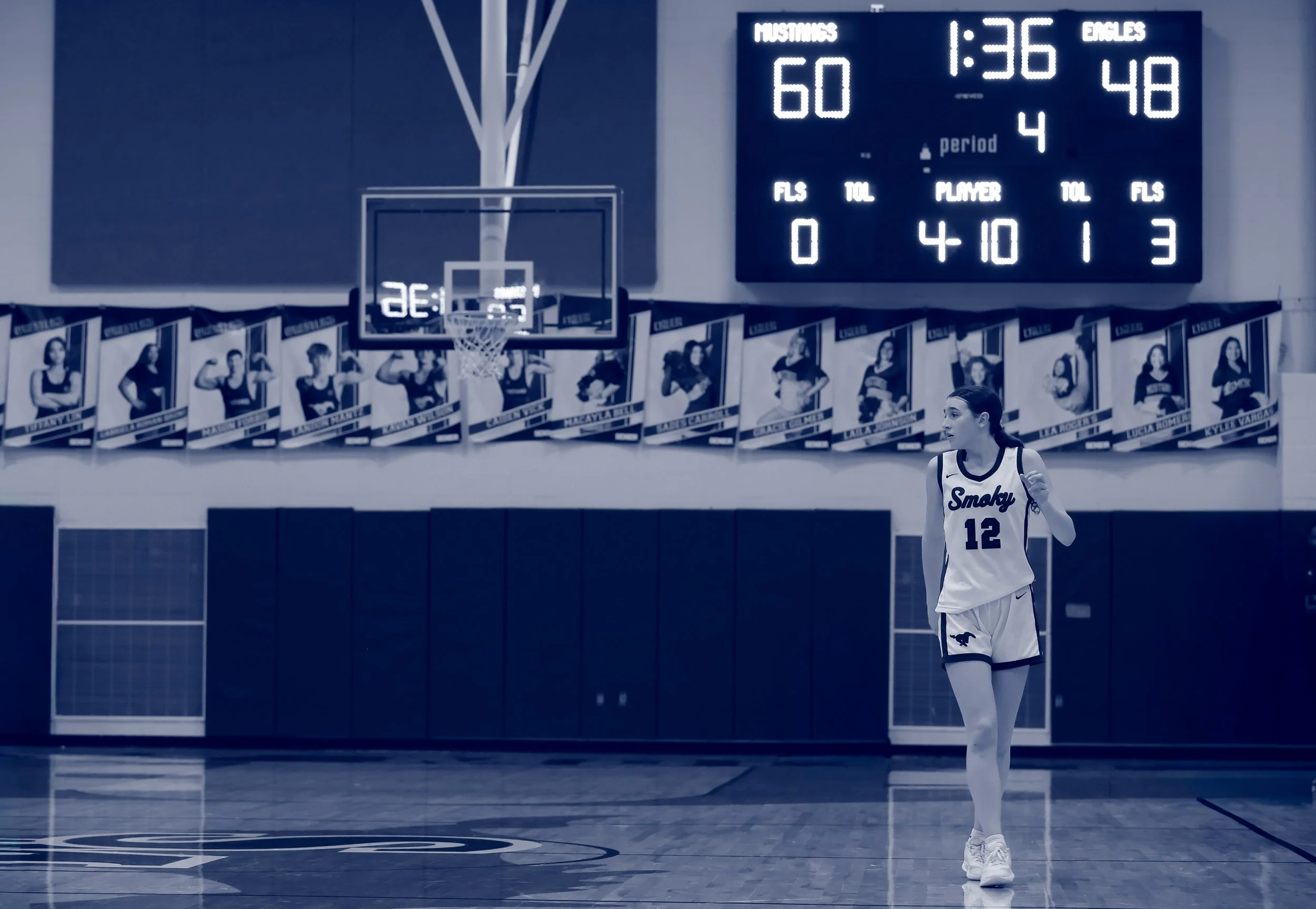 A female basketball player wearing a white uniform with the number 12 and the team name Smoky walking on a basketball court. A digital scoreboard displays the game statistics in the background.