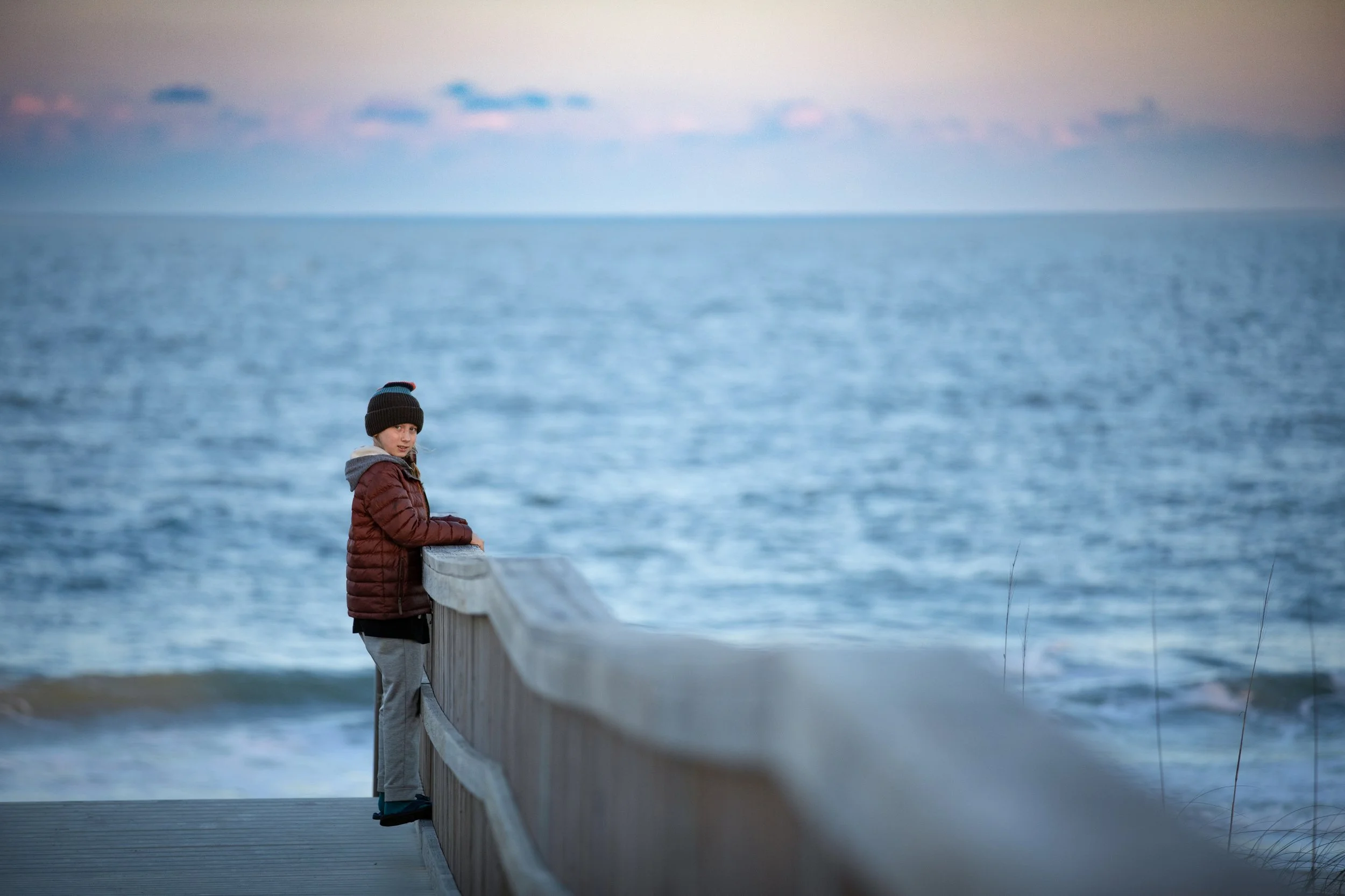 A young boy in a brown jacket and black beanie hat stands on a pier, leaning on the wooden railing, looking at the camera with the ocean and some clouds in the background.