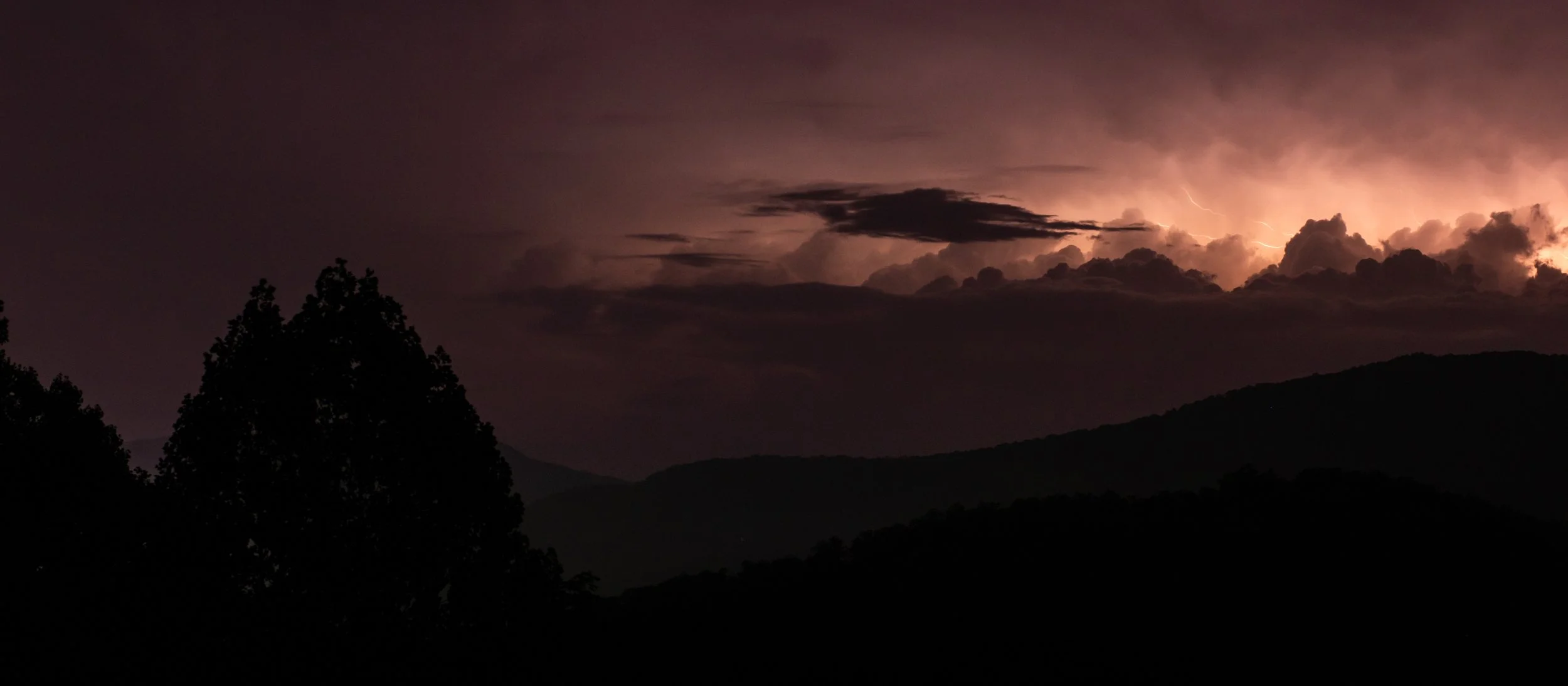 Nighttime scene of a storm with lightning illuminating dark clouds over mountains, with silhouetted trees in the foreground.