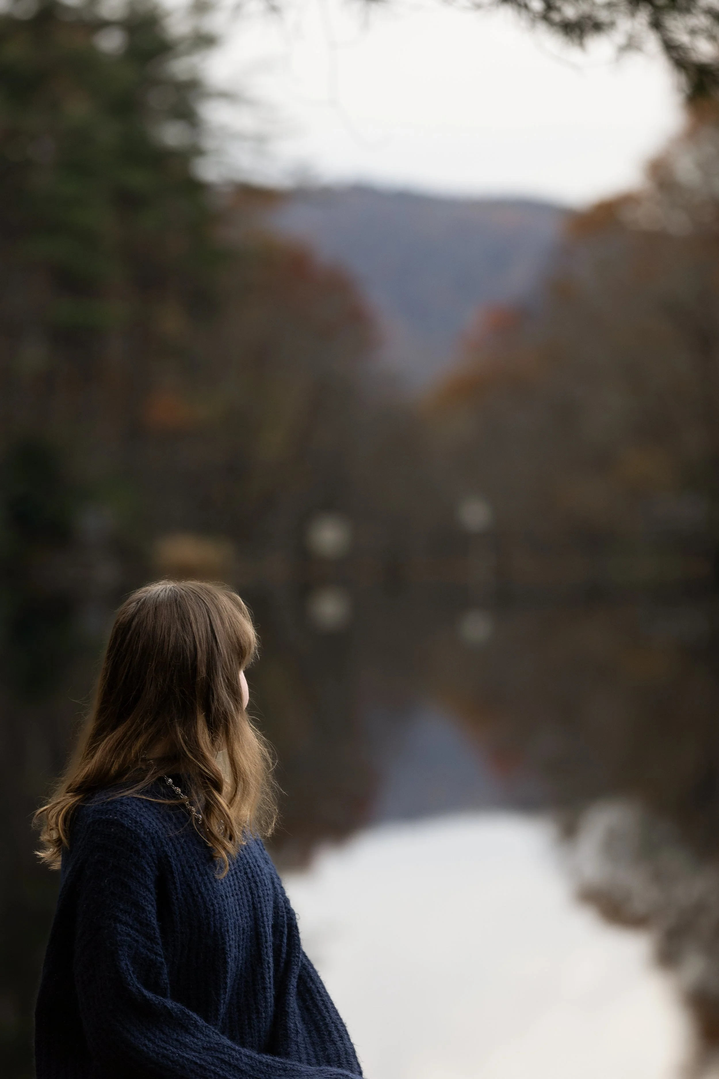 A woman with long brown hair, wearing a dark blue knitted sweater, is looking at a blurred outdoor scene with trees and a body of water in the background.