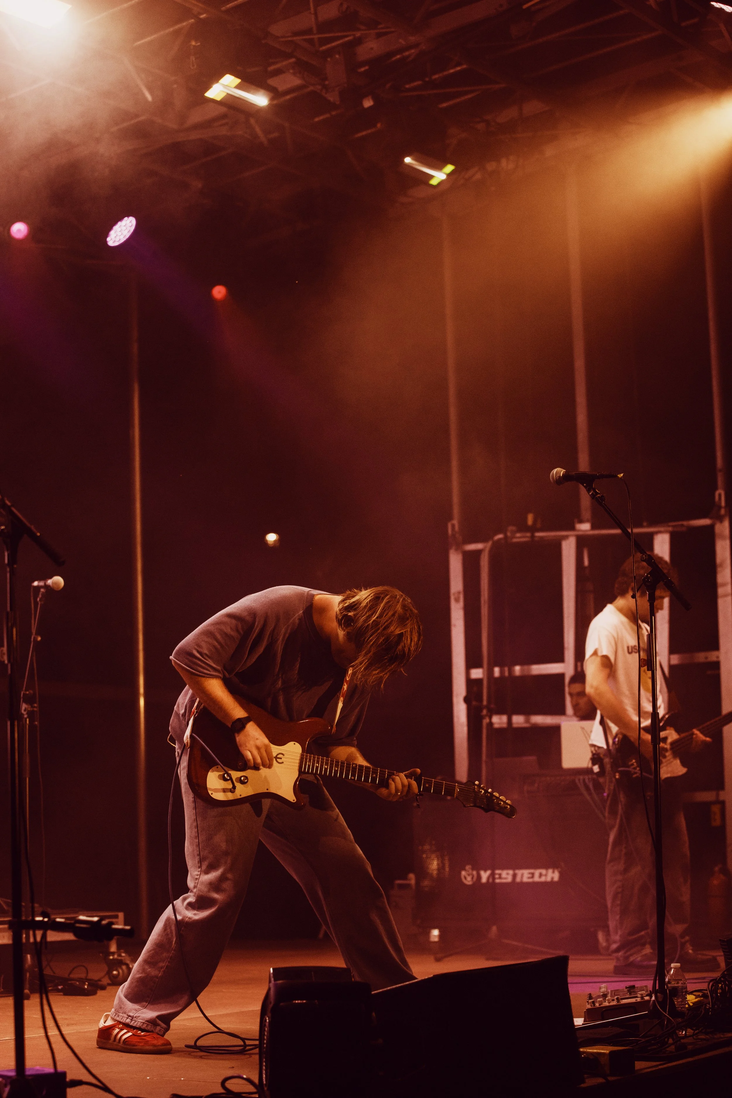 A musician playing an electric guitar on stage under colorful stage lights, with other band members and equipment visible in the background.
