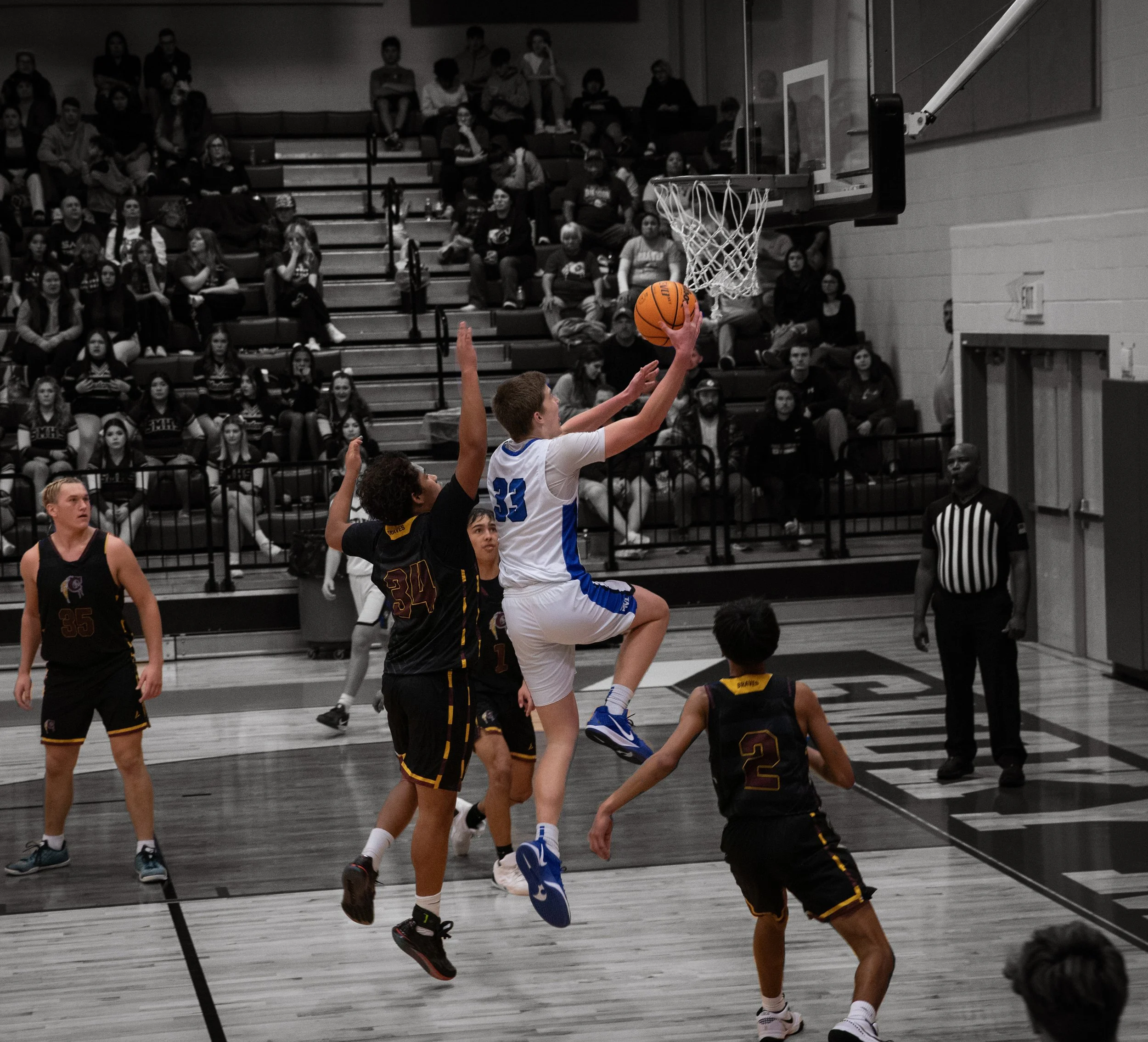 A basketball game with players in uniforms, one player in a white and blue jersey is jumping to shoot or dunk the ball, while another player in a black and yellow jersey is attempting to block. The game is taking place in an indoor gymnasium with spe