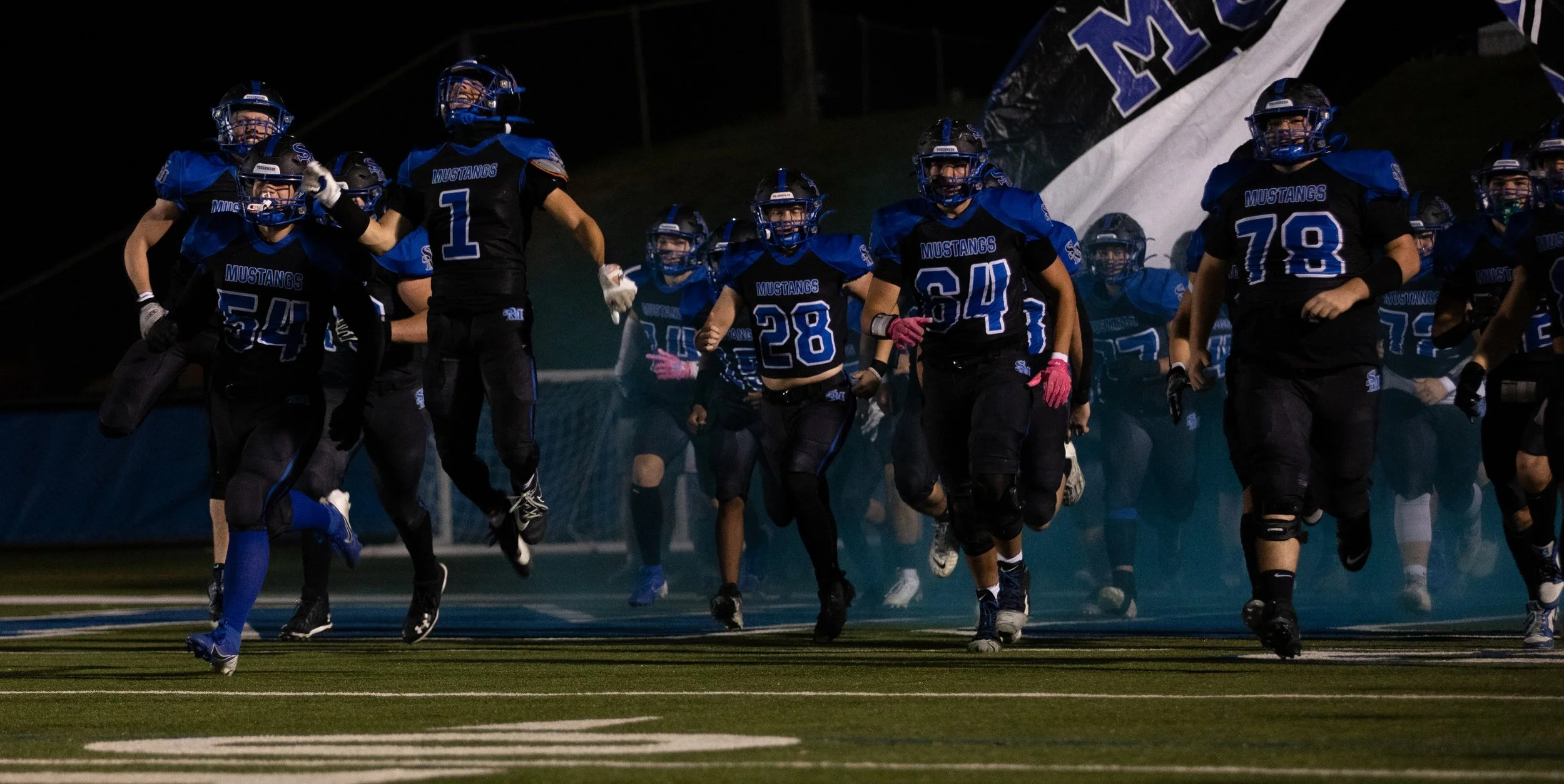 Youth football players in blue and black uniforms running onto the field during the night game.