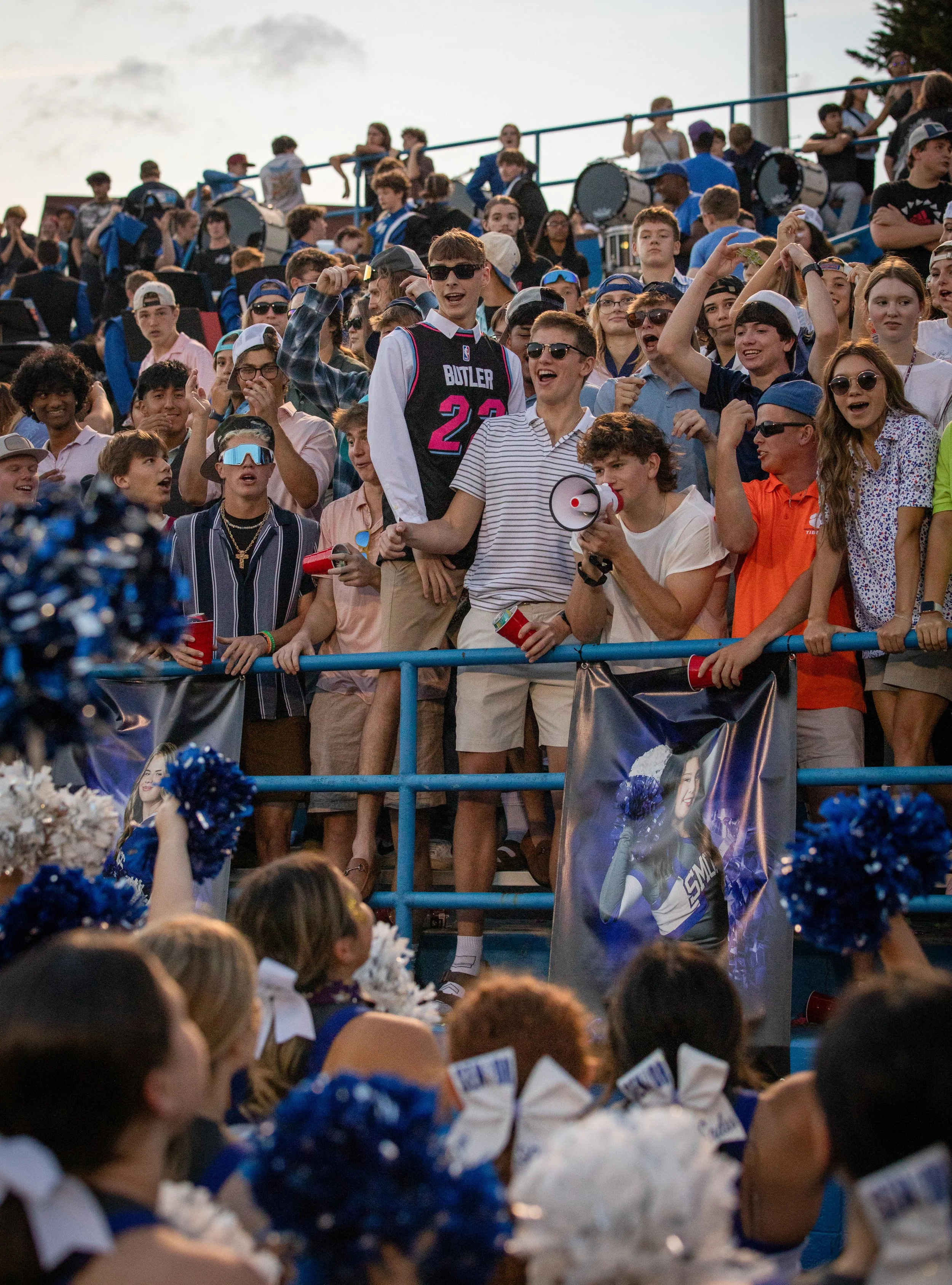 A large crowd of mostly teenagers at an outdoor event, some wearing sunglasses, hats, cheerleading uniforms, and holding pom-poms. There are banners and people in the background, with some holding cans and a few taking photos or videos.