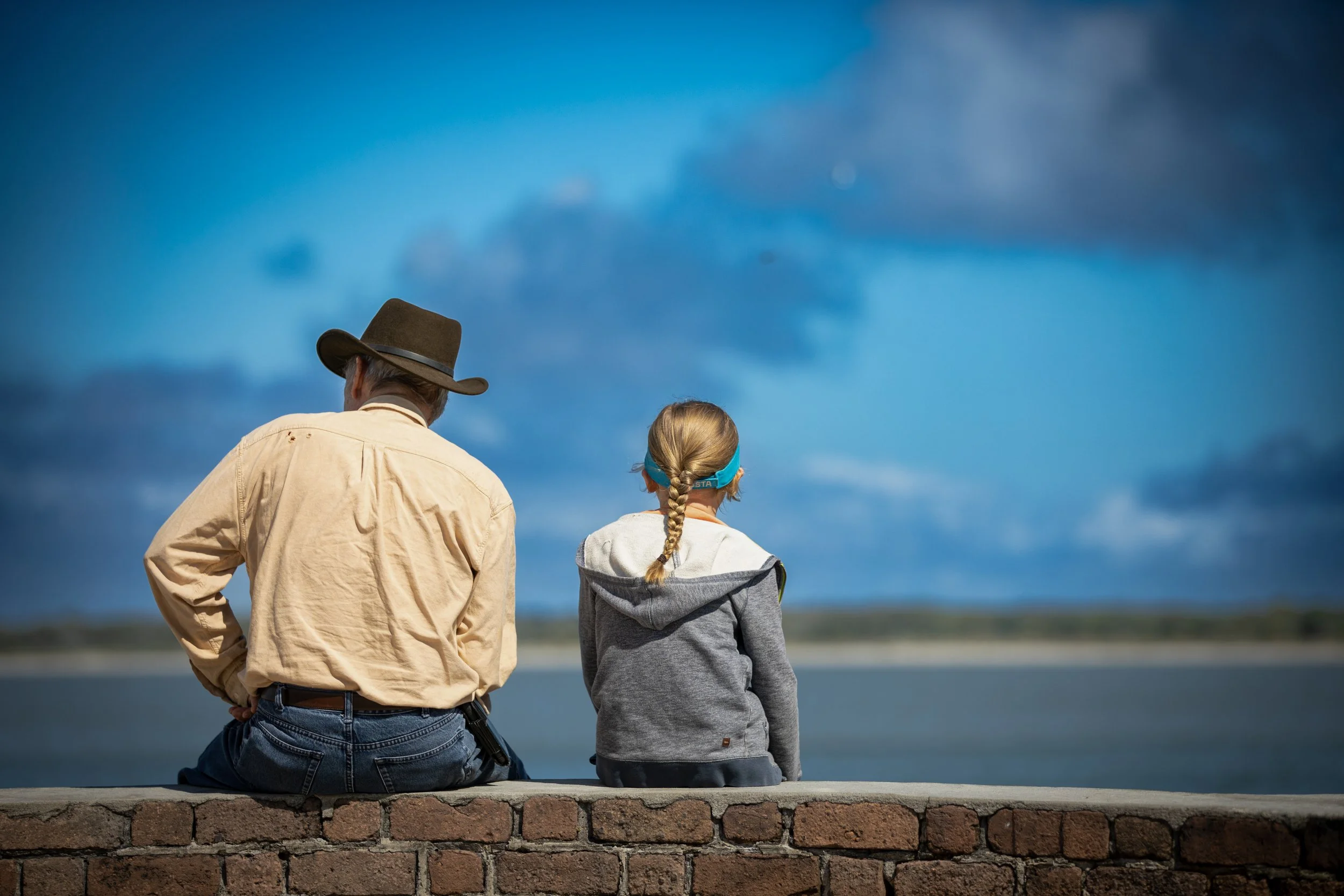 A man and a girl sitting on a brick ledge facing a body of water under a cloudy sky. The man is wearing a cowboy hat and beige shirt, while the girl has blond hair in a braid, wearing a gray hoodie and a blue headband.
