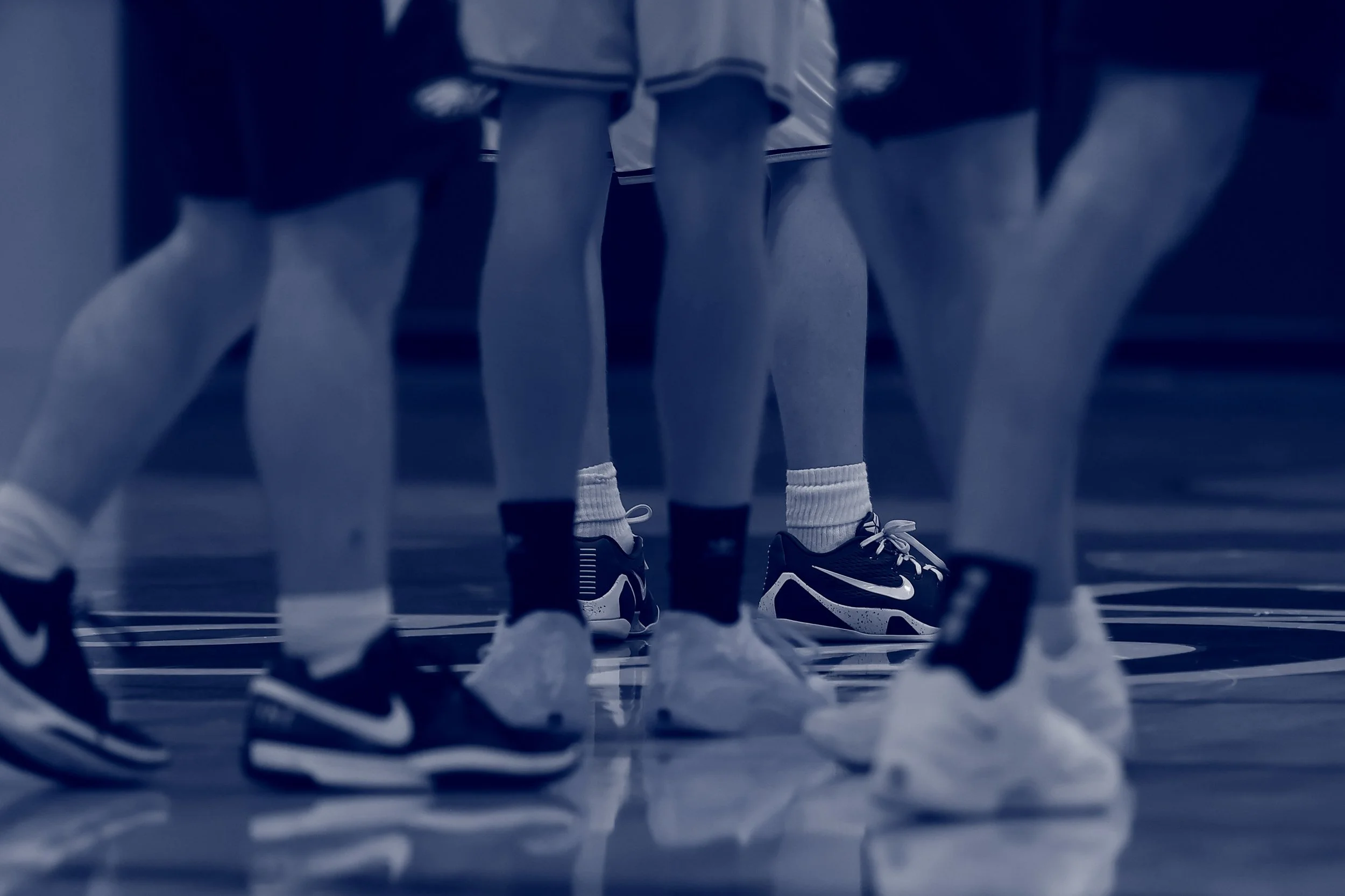 Close-up view of people standing on a sports court, focusing on their legs and feet, wearing athletic shoes and socks, with a striped sports jersey visible in the background.