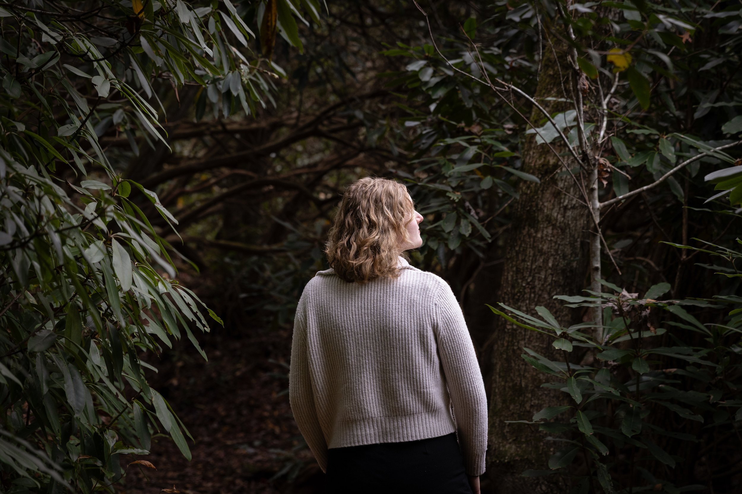 A person with curly hair and a beige sweater standing in a dense, dark forest surrounded by trees and green foliage.