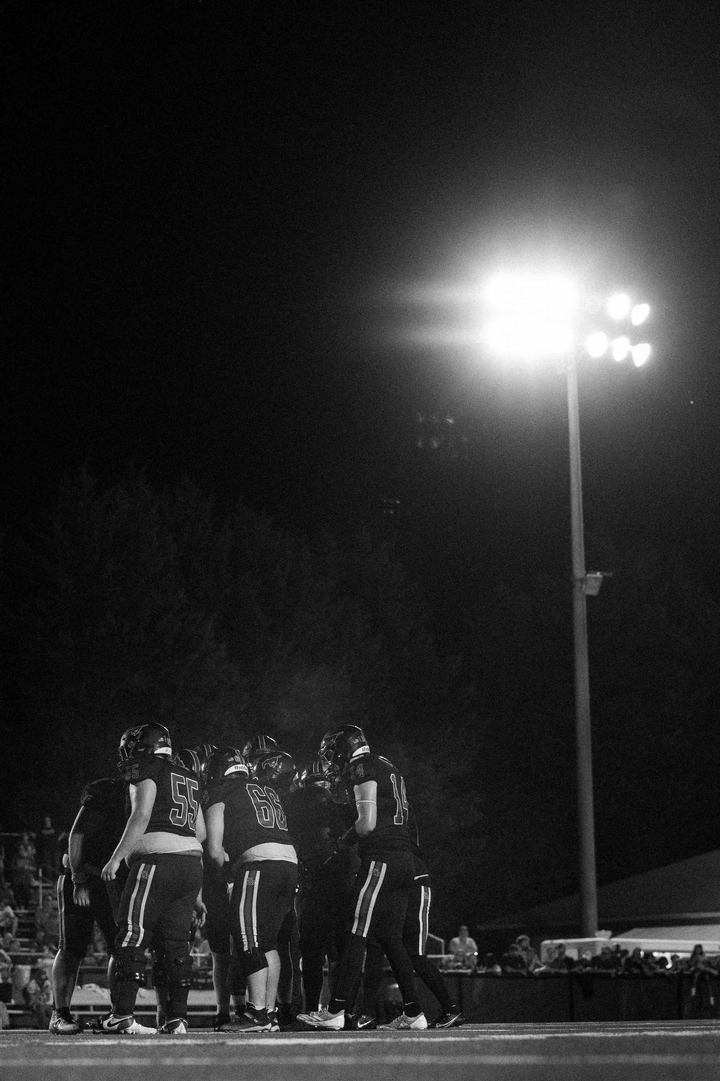 A group of football players in black uniforms huddle together on the field during a night game, with bright stadium lights in the background.