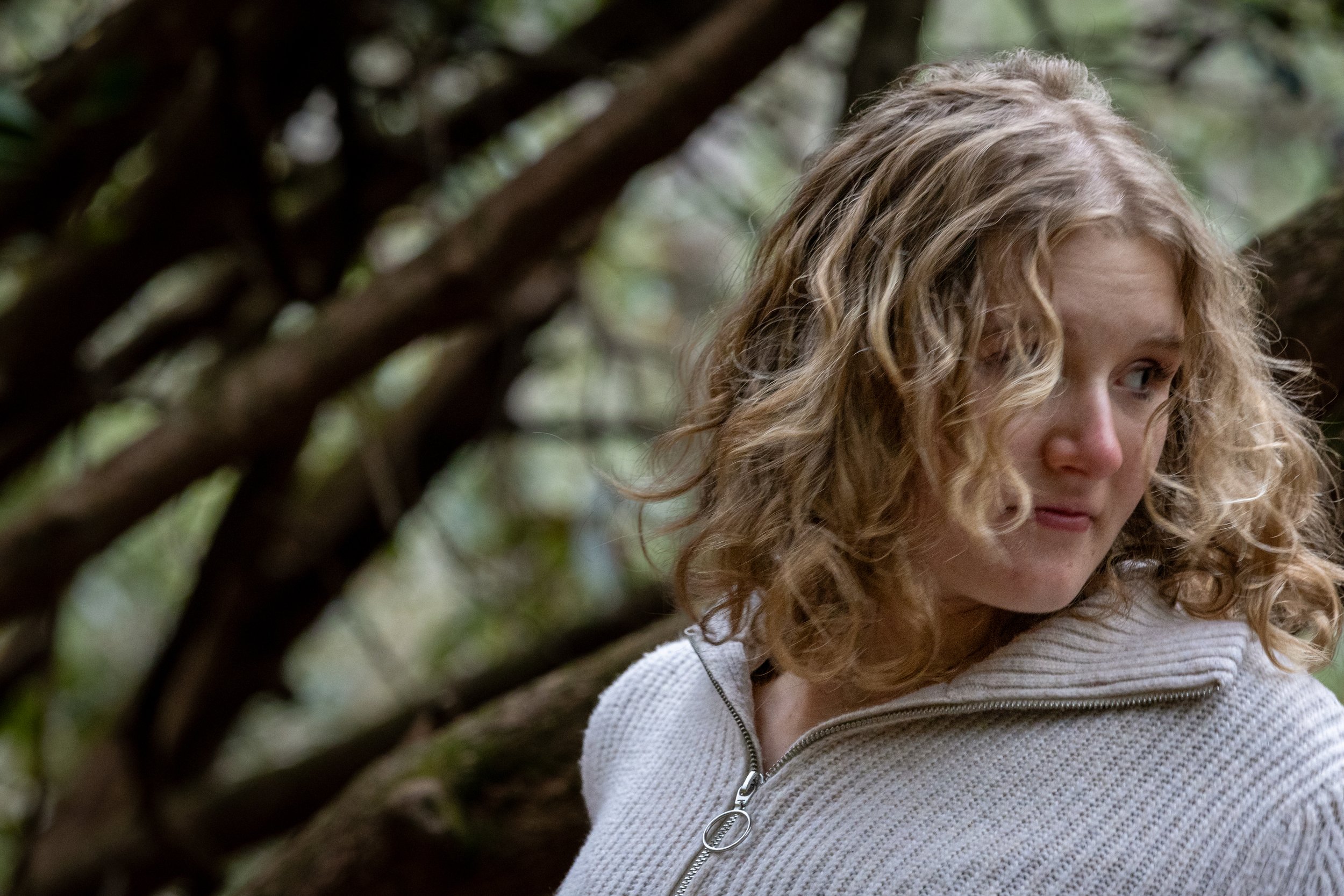 A woman with curly blonde hair wearing a white zip-up sweater outdoors in a wooded area.