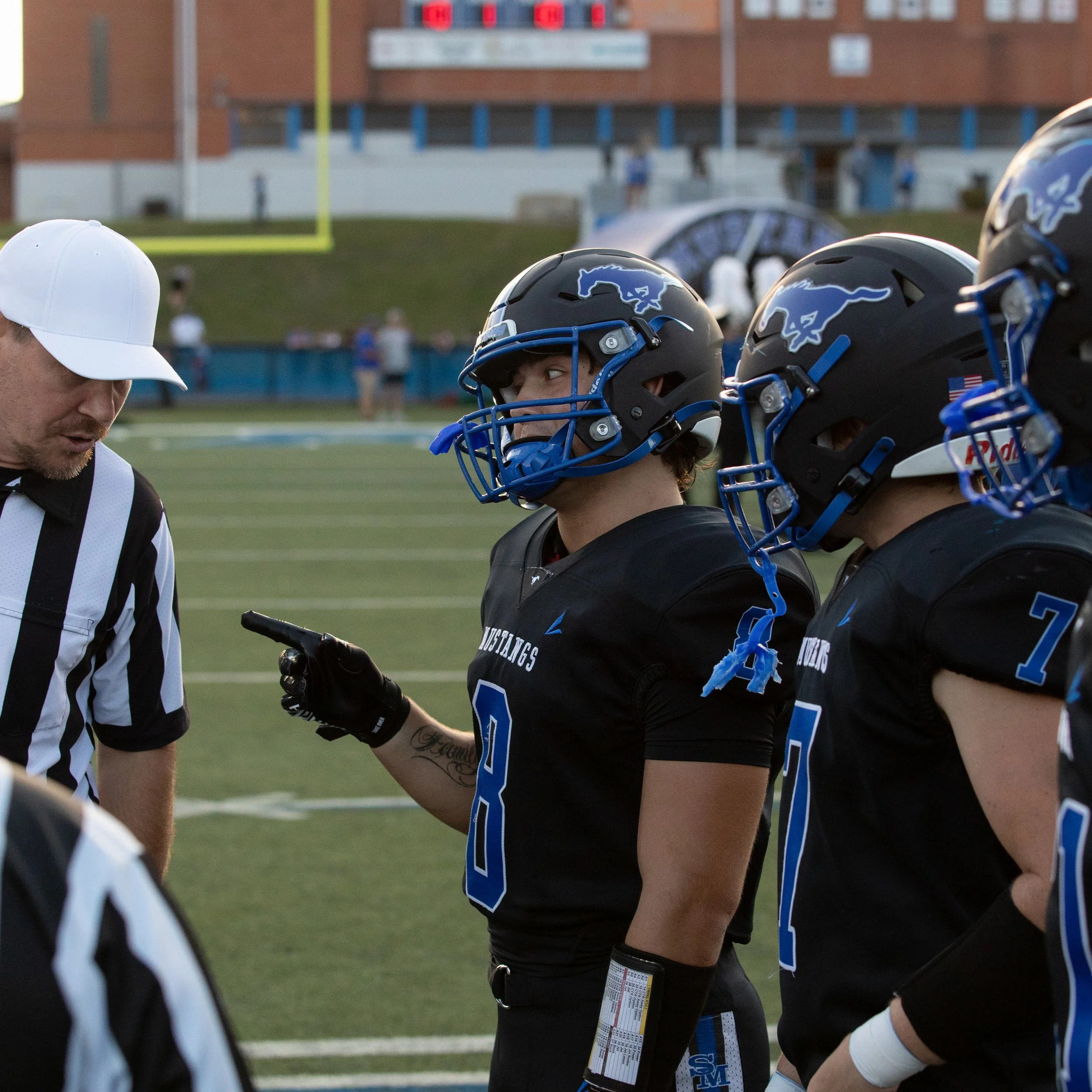 Close-up of high school football players wearing black and blue helmets with a mustang logo, standing in a huddle on a football field with a referee and blurred background of a school building and scoreboard.