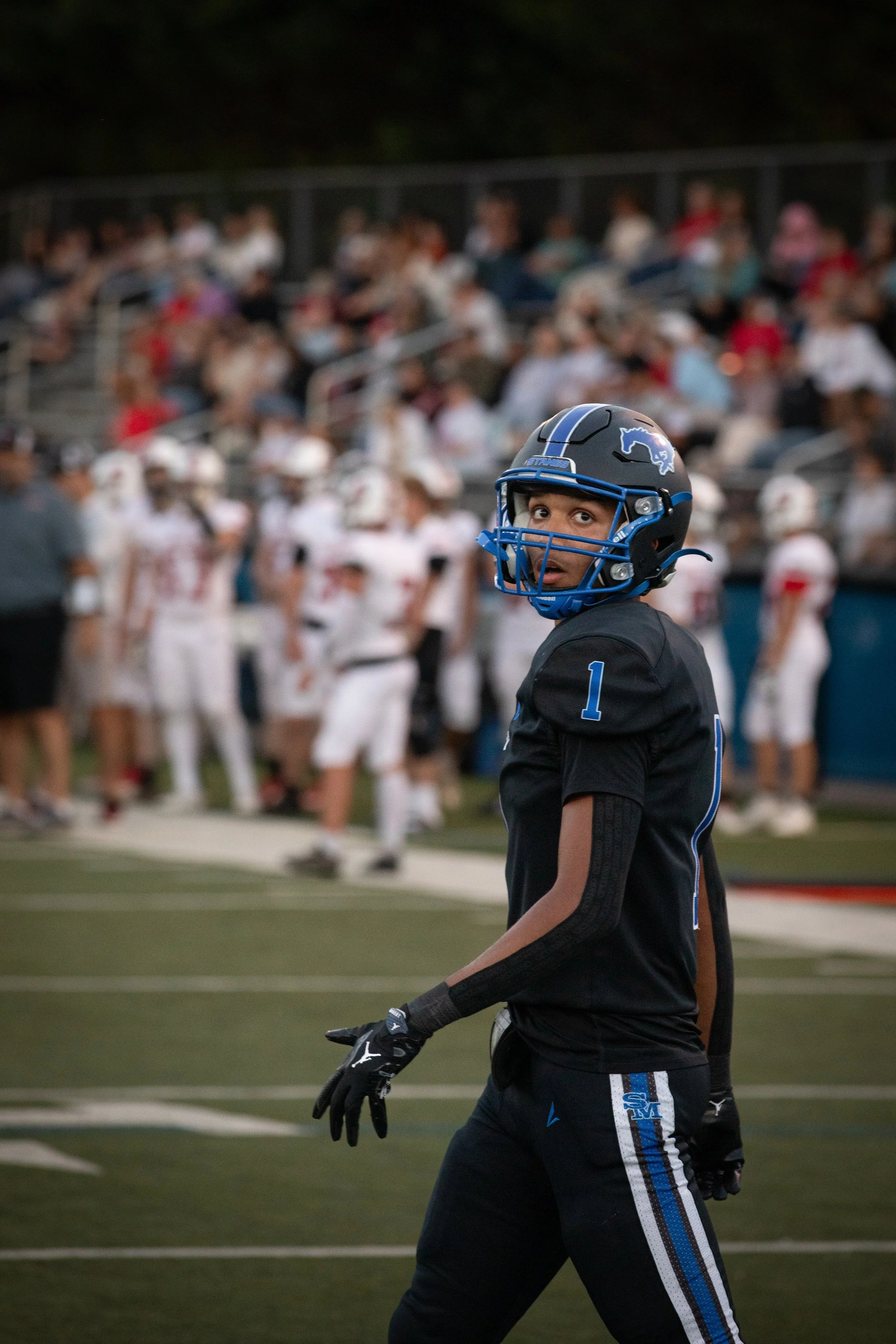 A football player wearing a black uniform and a helmet with a blue face mask and the number 1 on the shorts stands on a football field with a crowd of spectators in the background.