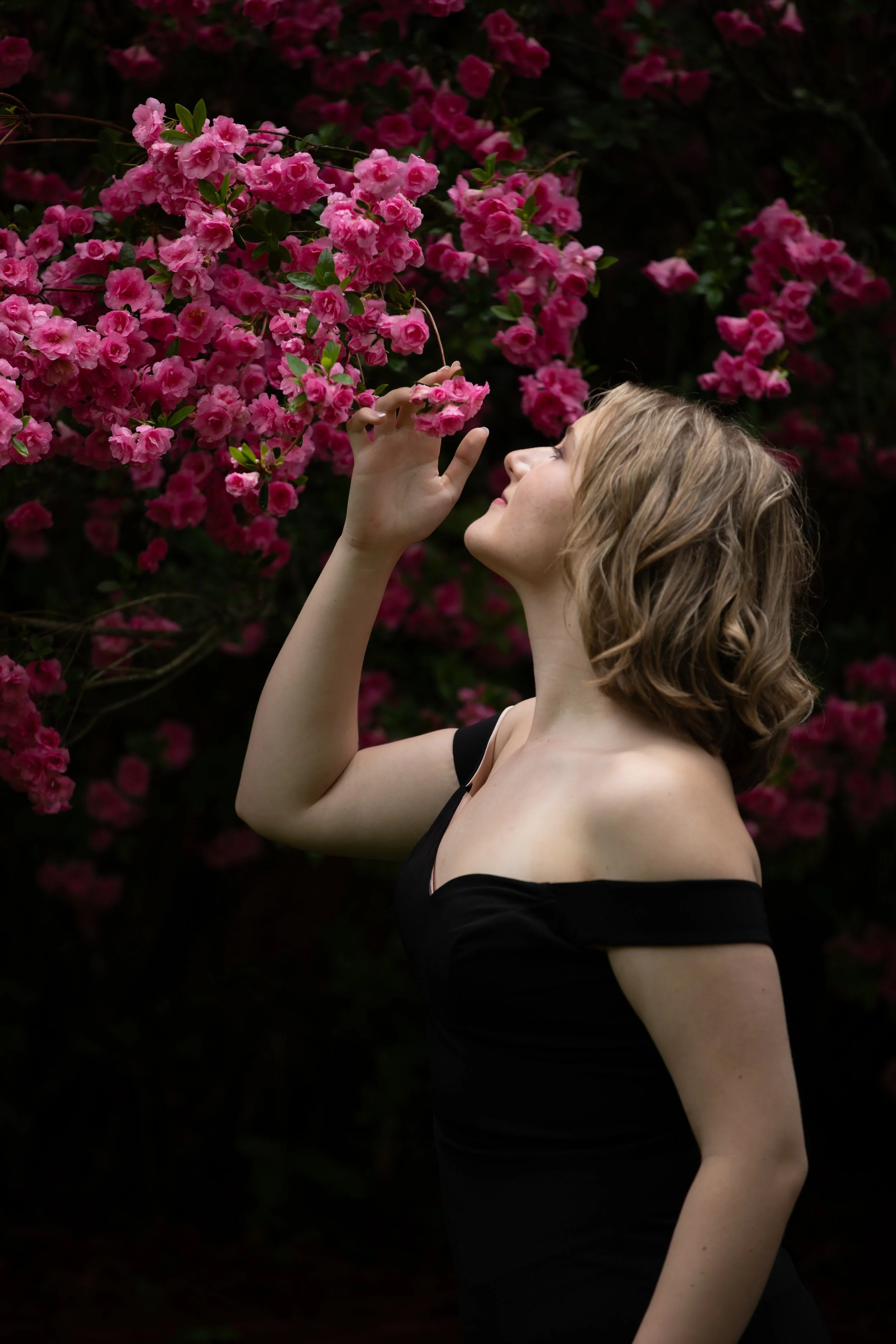 A woman with short, wavy blonde hair is standing outdoors next to a pink flowering bush, touching one of the flowers with her finger while looking at it.