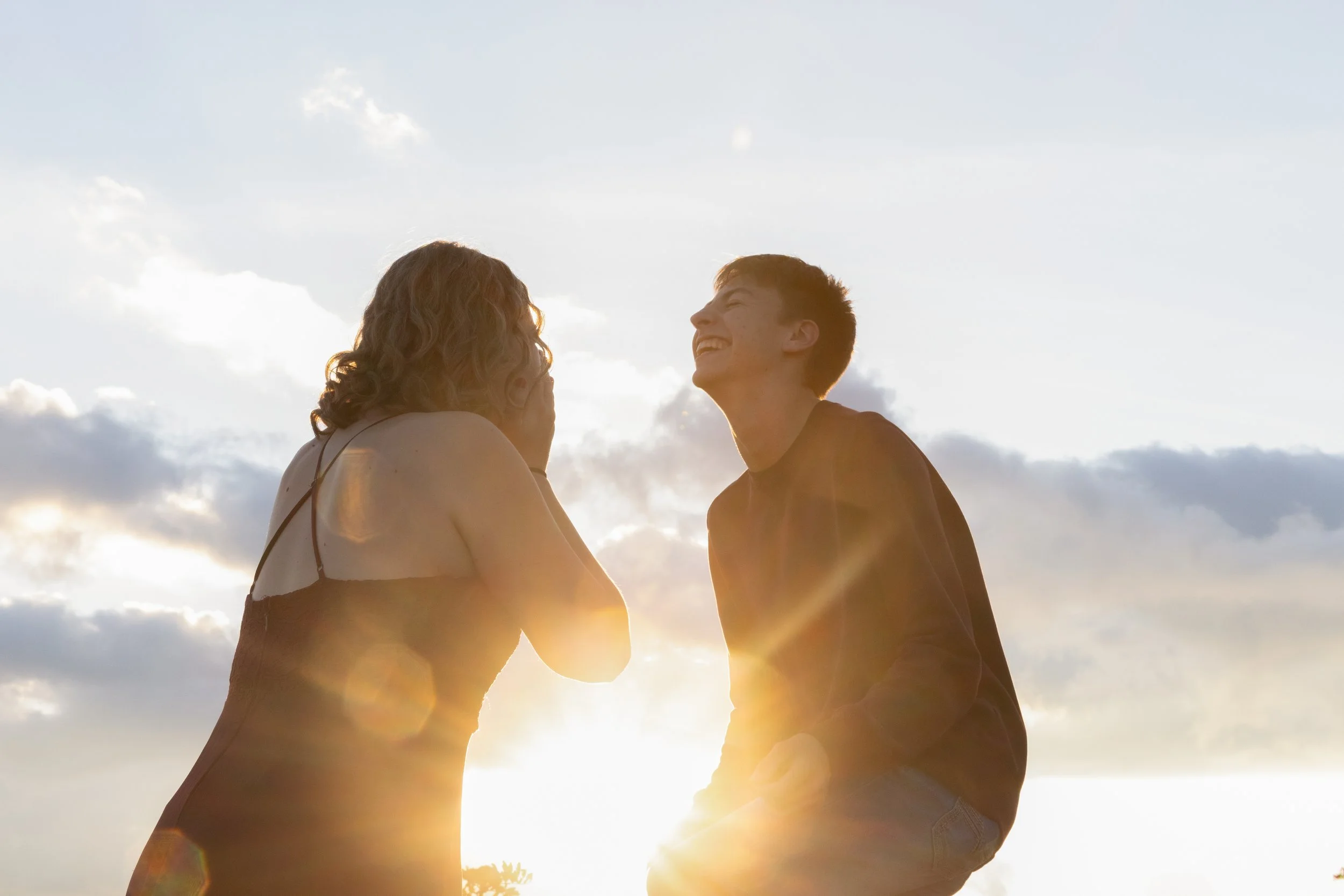 A couple is outdoors during sunset, laughing and facing each other, with sunlight creating a warm glow and lens flare effect.