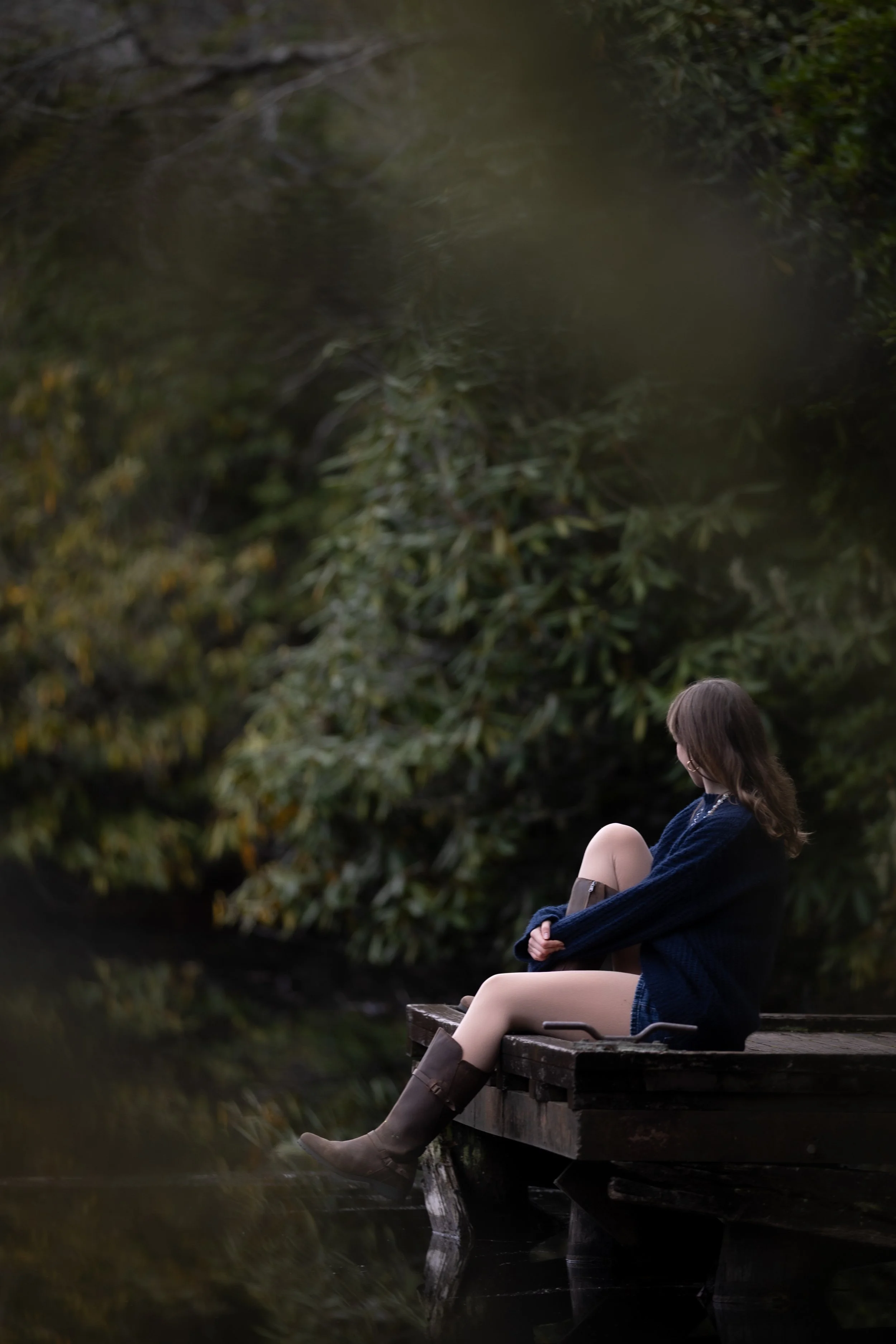 A young woman sitting on a wooden dock by a pond, wearing a navy blue sweater, beige shorts, and knee-high boots, with greenery in the background.