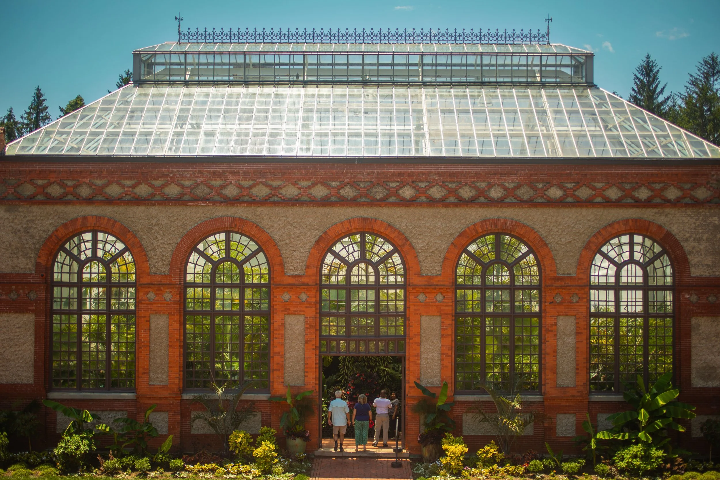 A historic brick greenhouse with large arched windows and a glass roof, surrounded by lush greenery and flowers, with three people walking towards the entrance.