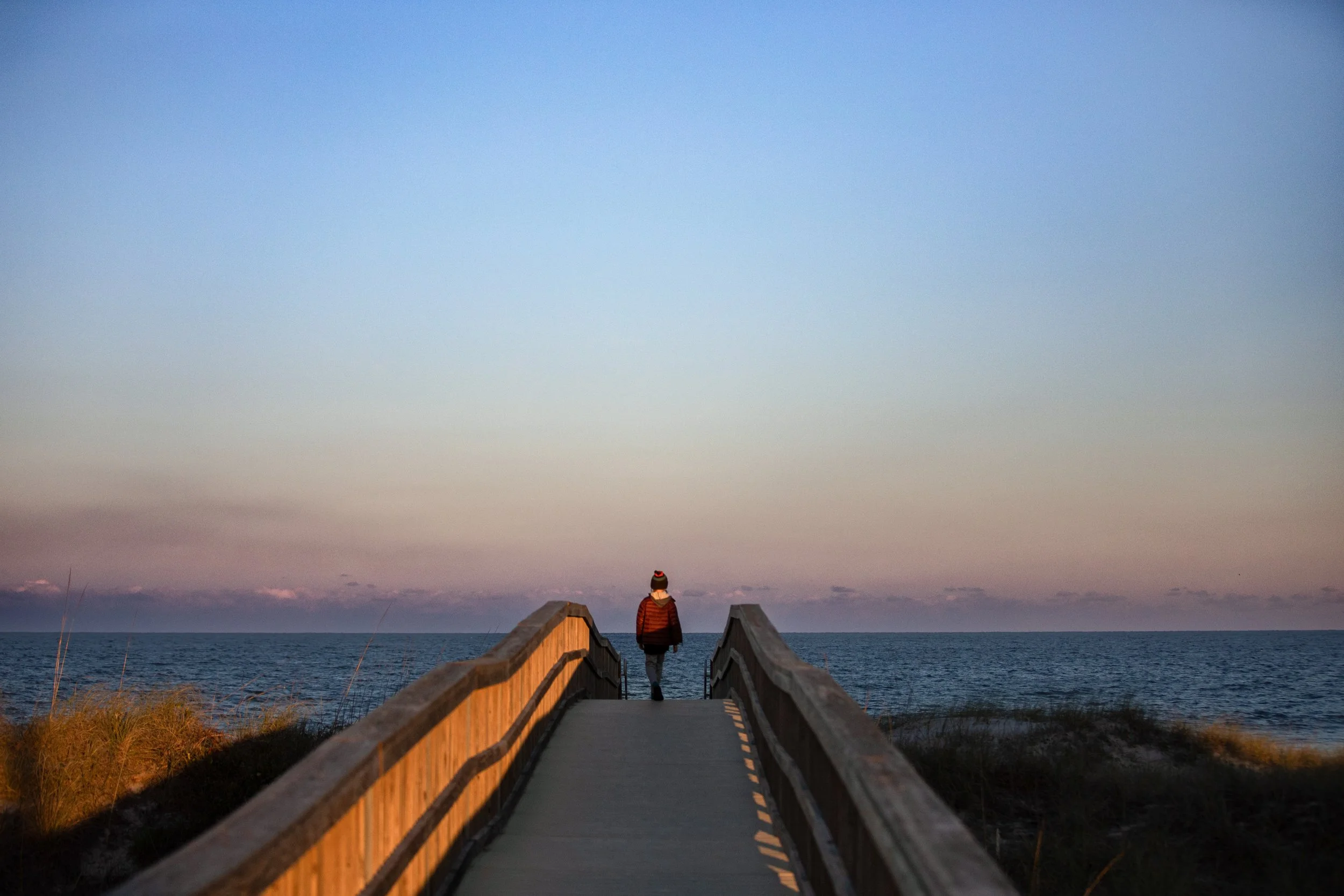 Person walking down a wooden pier towards the ocean at dusk or dawn with a colorful sky.