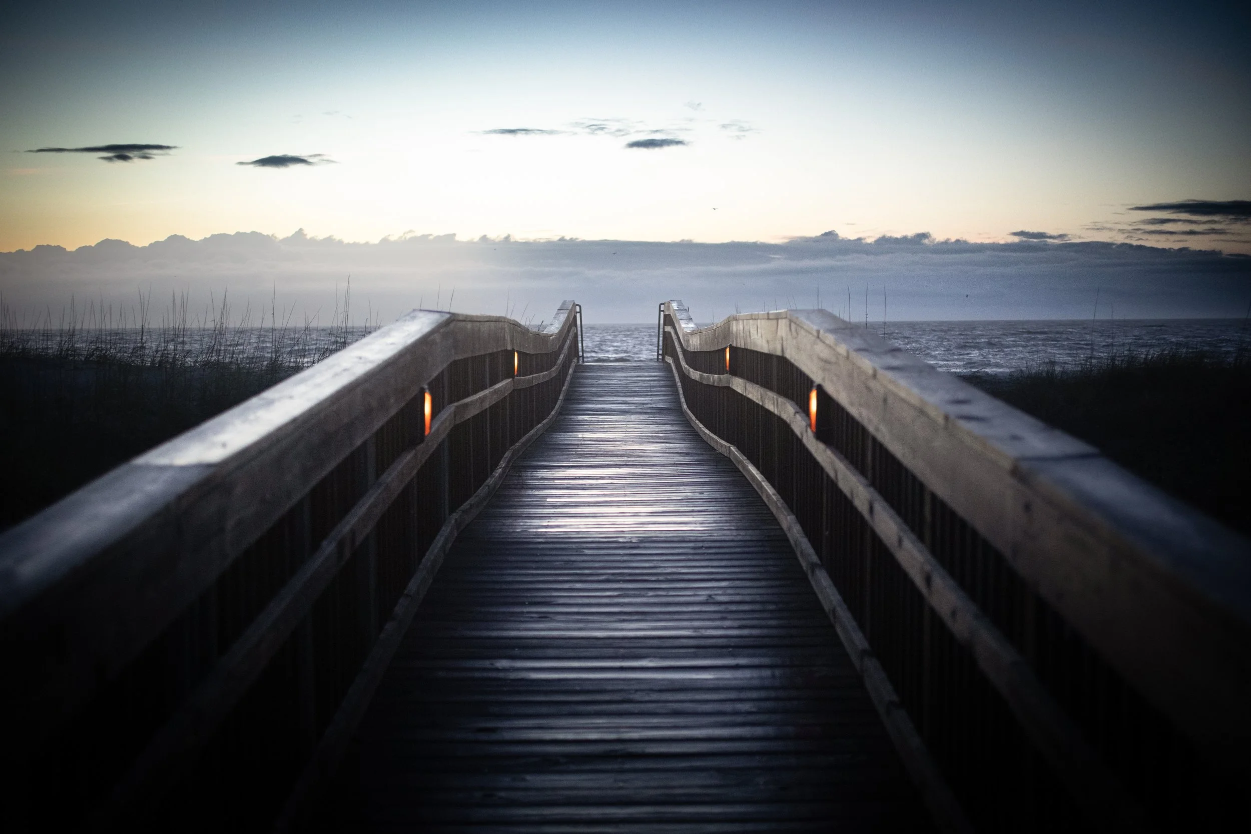 A wooden pathway leading to the beach at dusk, with the ocean in the background and a cloudy sky overhead.