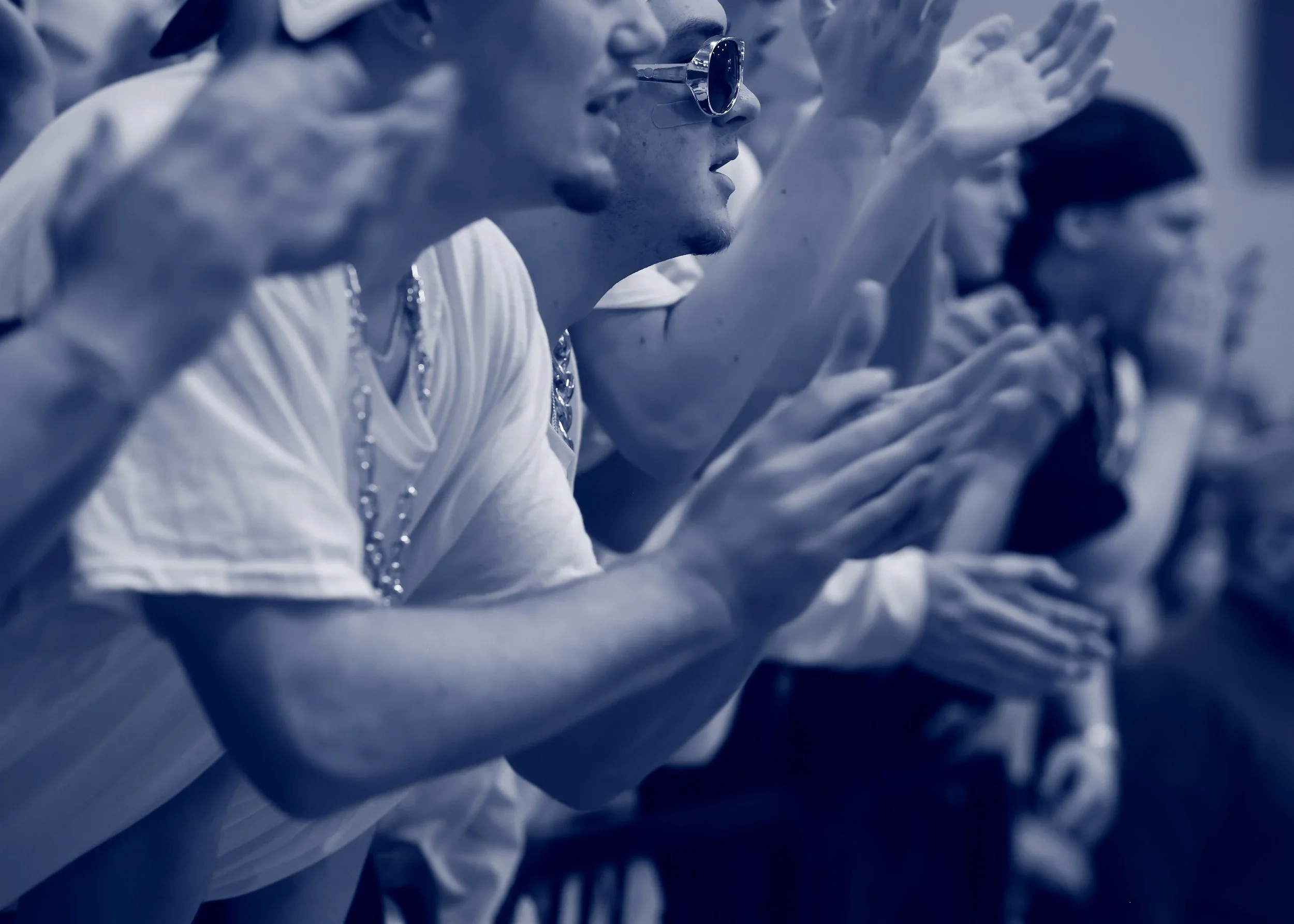 Close-up of a person wearing sunglasses and a chain necklace, with their hand raised near their face, in a stylized, monochromatic blue toned image.