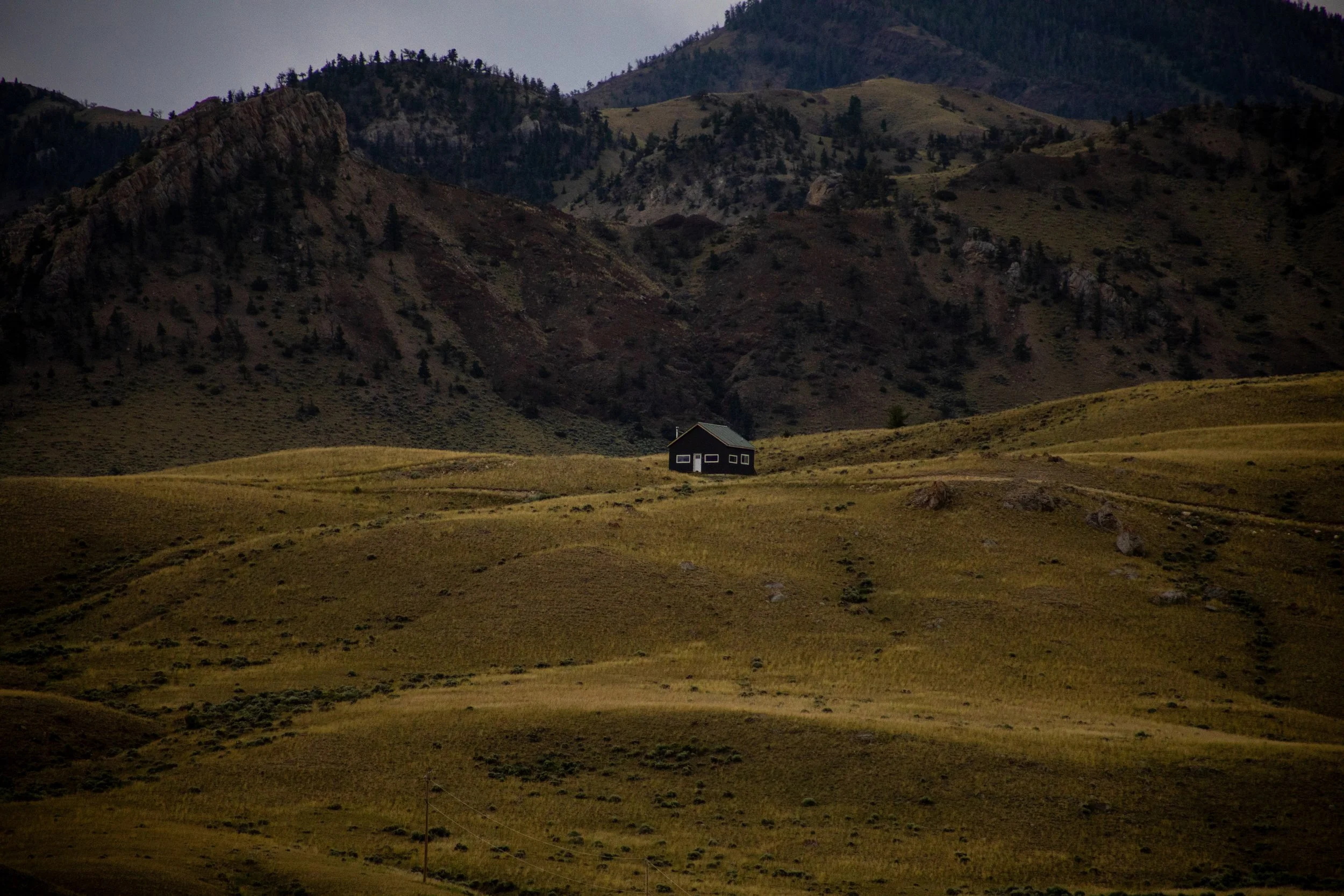 A small house with a dark exterior and white window frames sits in a grassy field at the foot of mountain ranges with rocky slopes and sparse trees.