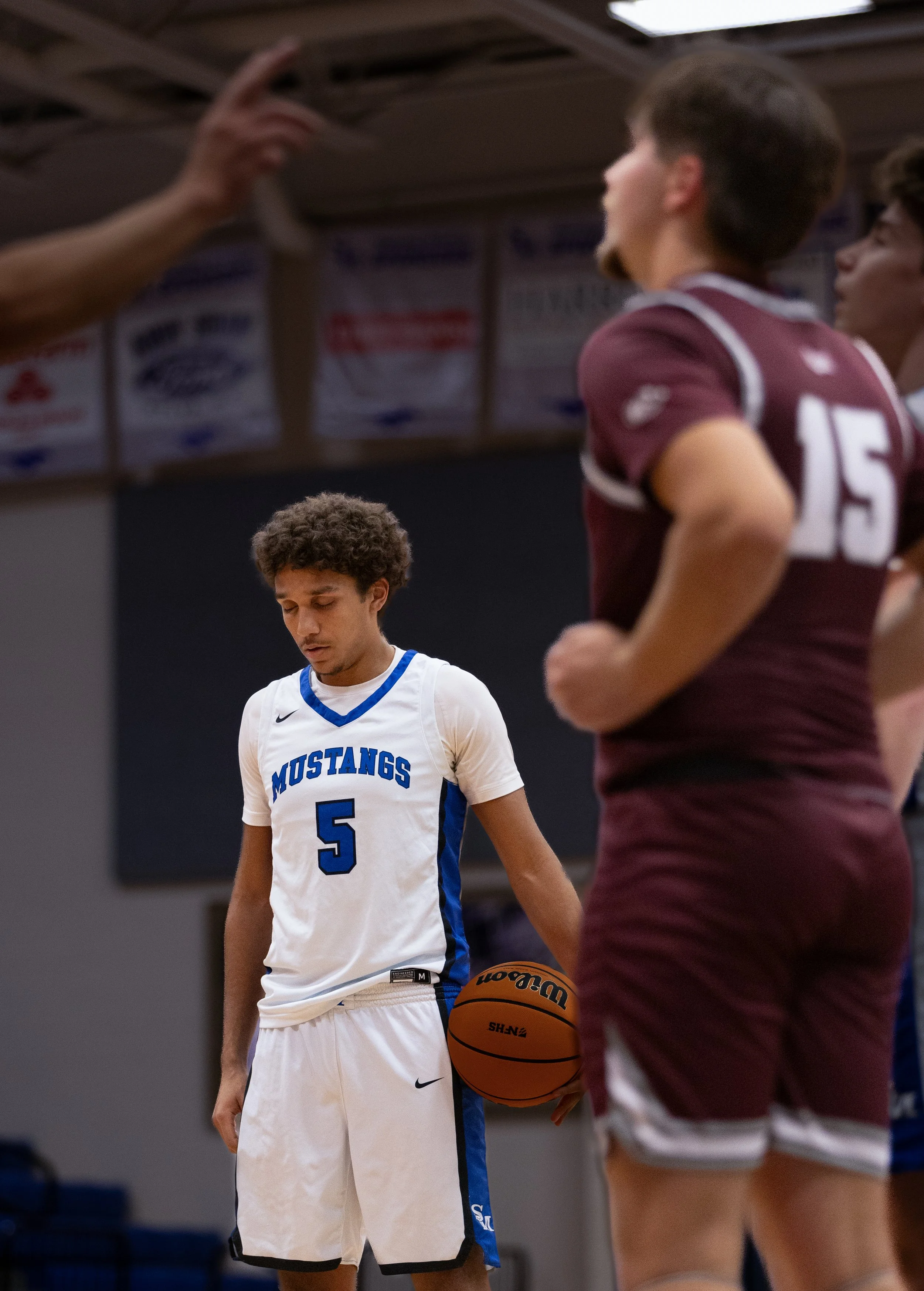 A basketball player in a white jersey with blue accents and the number 5 is standing with his eyes closed. Opposite, a player in a maroon jersey with the number 15 and maroon shorts holds a basketball close to her body, partially visible, with her ar