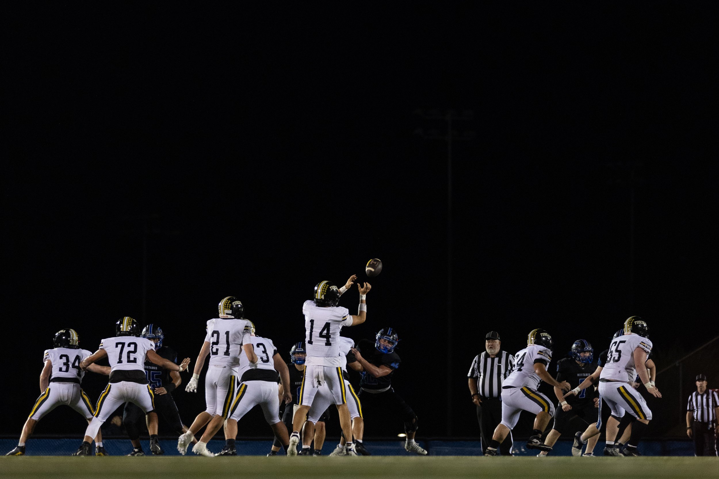 American football game at night with players from two teams lined up at the line of scrimmage. Player number 14 is about to throw the football, with a referee standing nearby. The dark sky and stadium lights illuminate the scene.