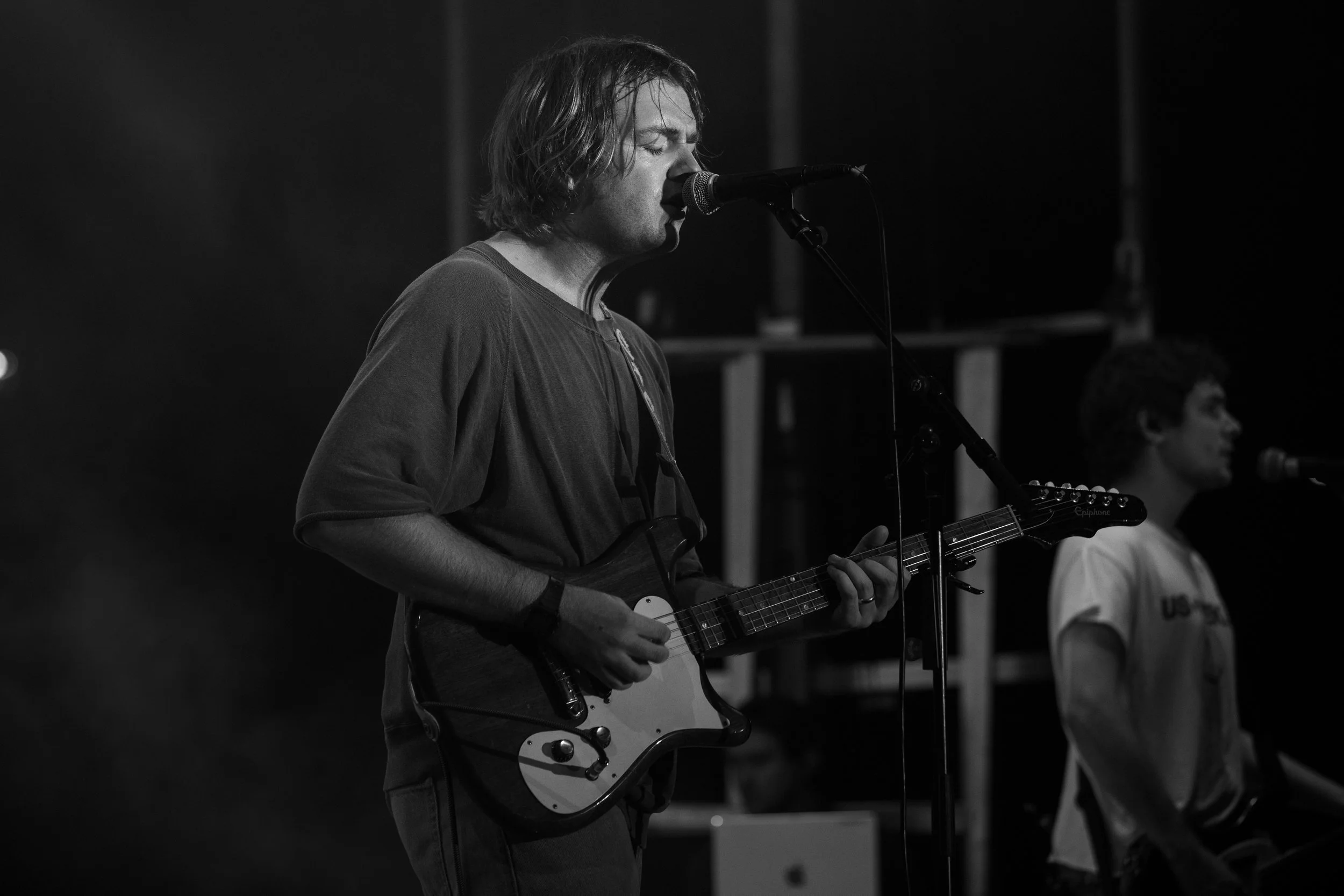 A person with shoulder-length hair singing into a microphone while playing an electric guitar in a dark setting.