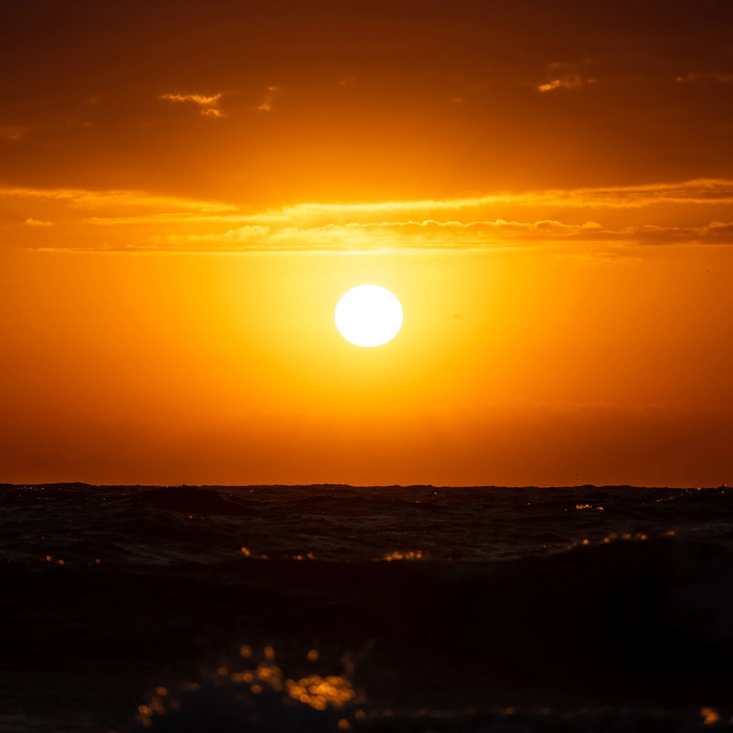 A sunset over the ocean with the sun near the horizon, casting an orange glow across the sky and water.