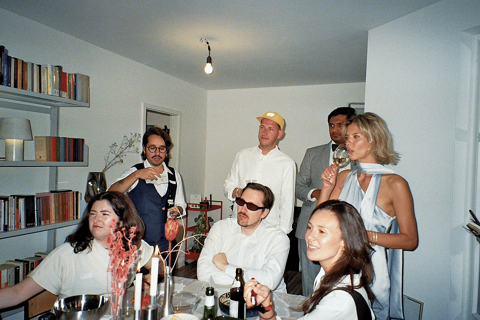 People at a house gathering, sitting and standing around a table with food and drinks, in a living room with bookshelves.
