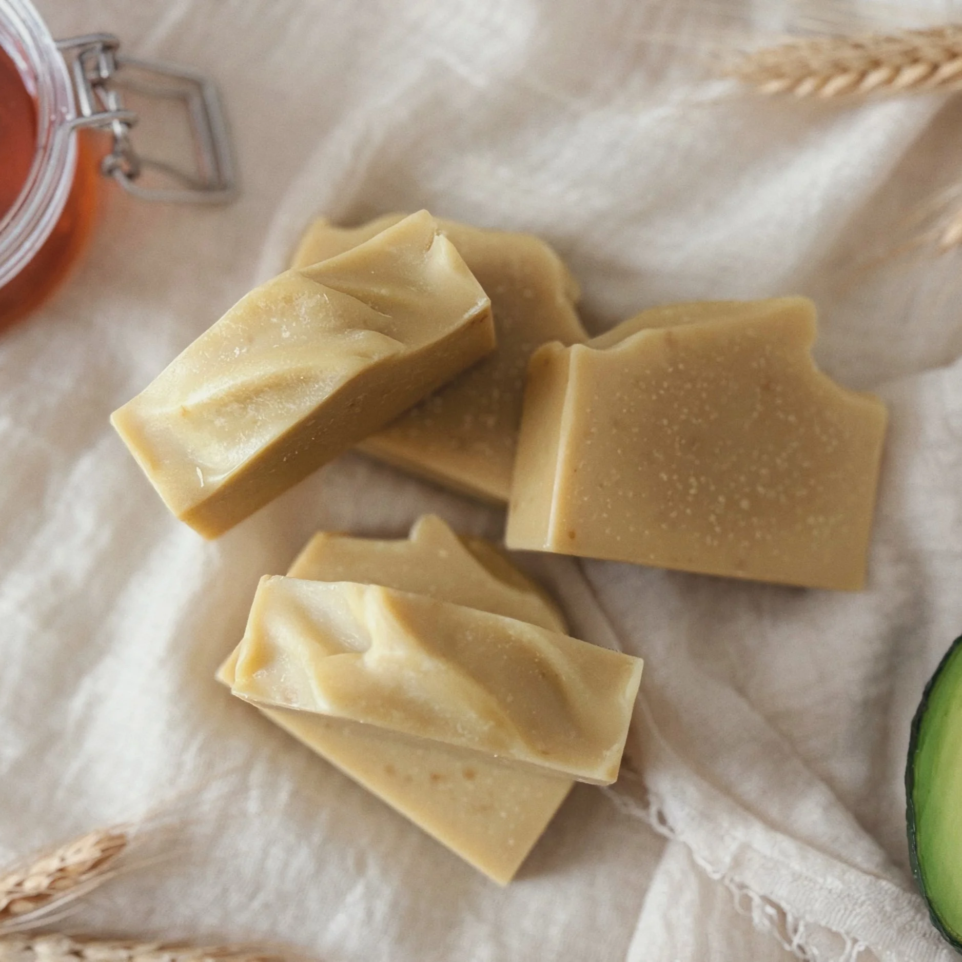Close-up of several pieces of beige-colored soap on a beige cloth, with a jar of orange-colored substance and a wheat stalk in the background.