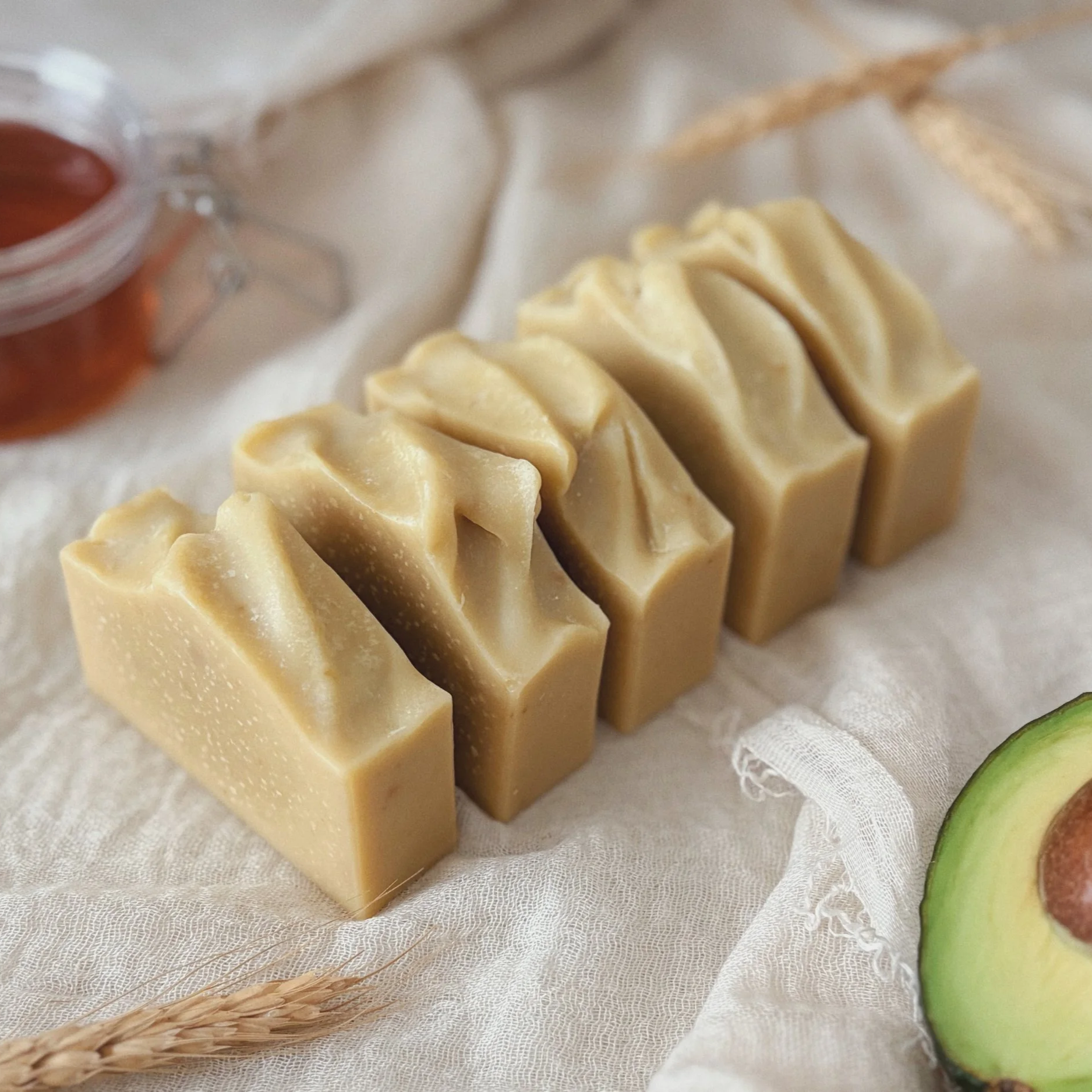 Close-up of a row of creamy beige artisan soap bars on white cloth, with wheat stalks and sliced green fruit in the background.