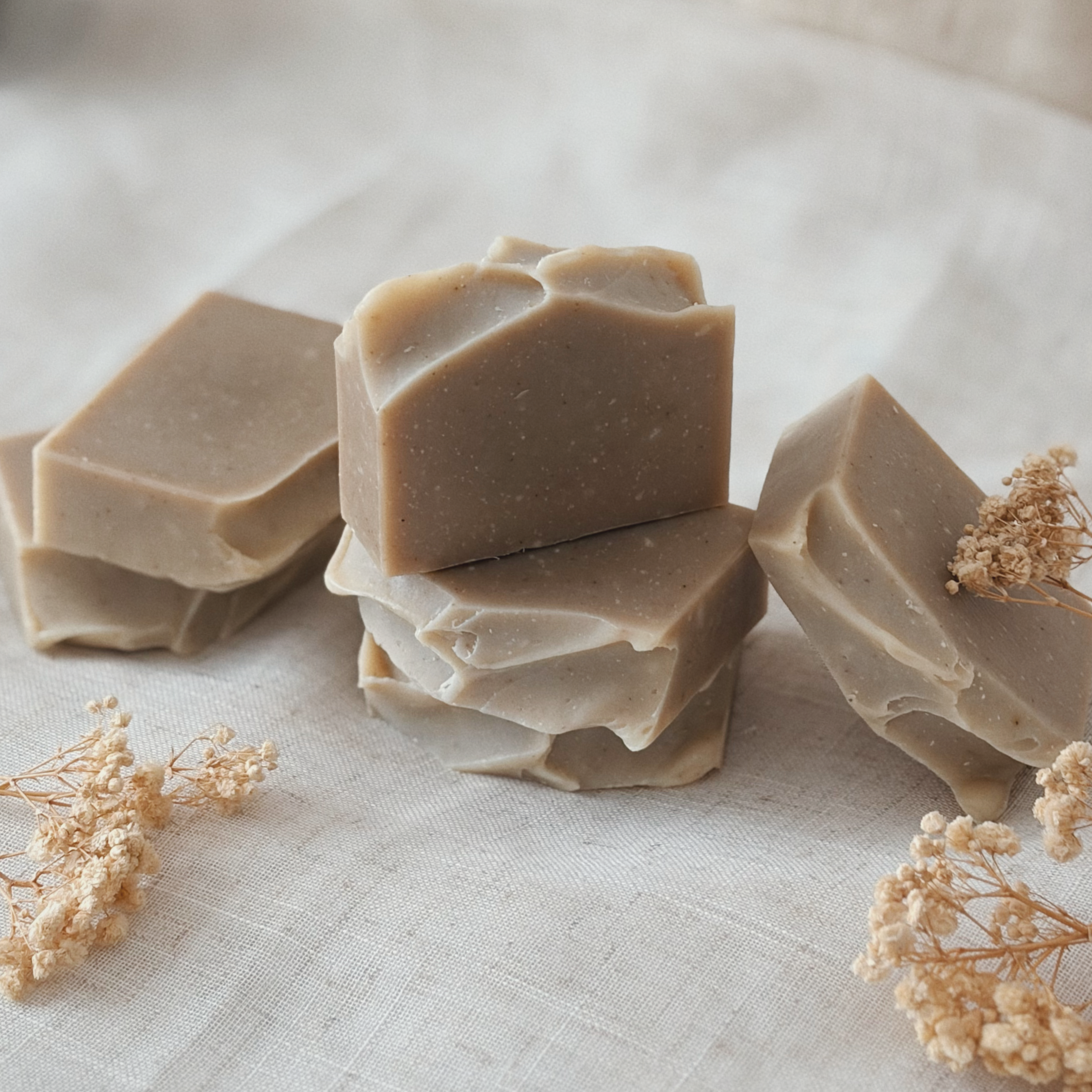 A close-up of several beige-colored soap bars with angular edges, arranged on a white surface with dried flowers nearby.
