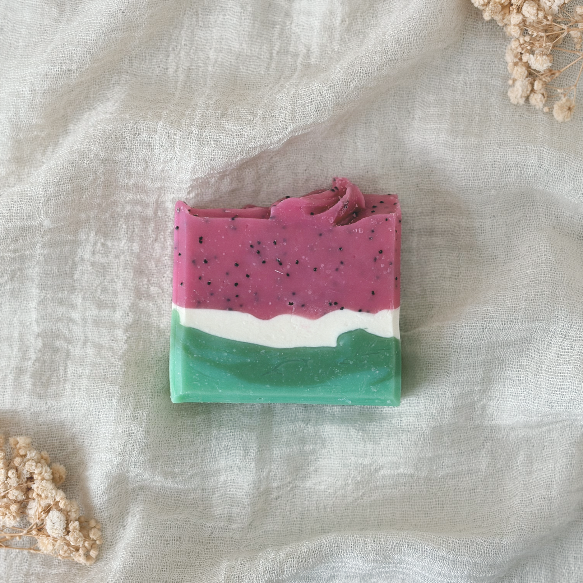 Close-up of a layered handmade soap bar with pink, white, and green colors, adorned with black specks, resting on a white textured fabric with dried flowers nearby.