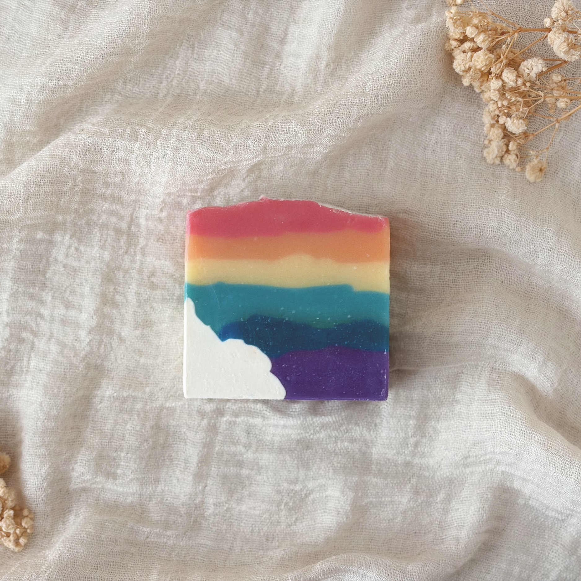 A colorful, striped soap bar with shades of pink, orange, yellow, green, blue, purple, and white resting on a light-colored fabric surface with dried flowers nearby.
