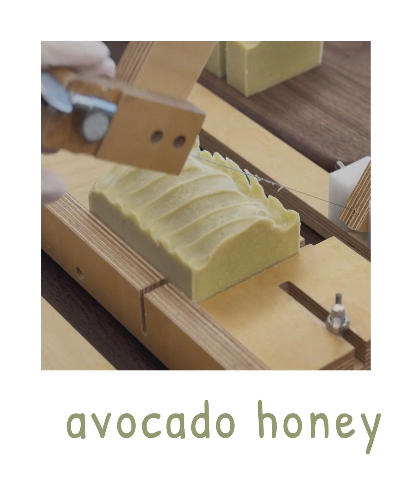 Close-up of soapmaking process with a wooden frame, yellow soap in a mold, and a person holding a metal spoon over the soap. The text 'avocado honey' is visible at the bottom.