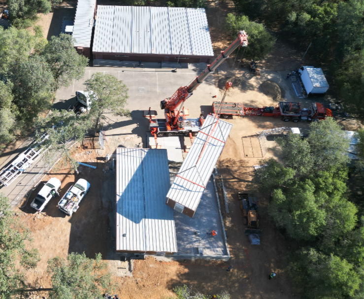 Construction site with a crane lifting a metal roof panel onto a building, surrounded by trees and construction vehicles.