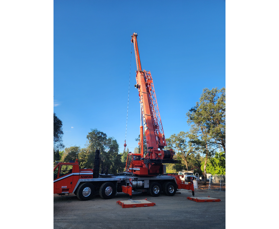 A large orange crane truck with extended boom working outdoors on a clear day, surrounded by trees and parked cars.