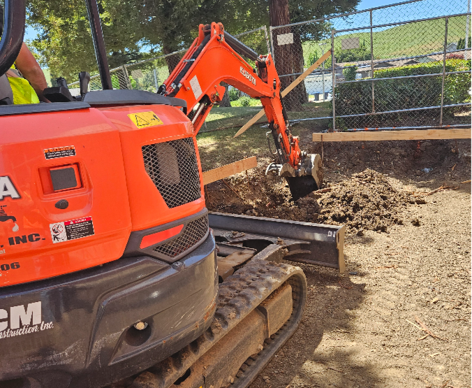 A small orange Kubota excavator digging into dirt on a construction site near a chain link fence.