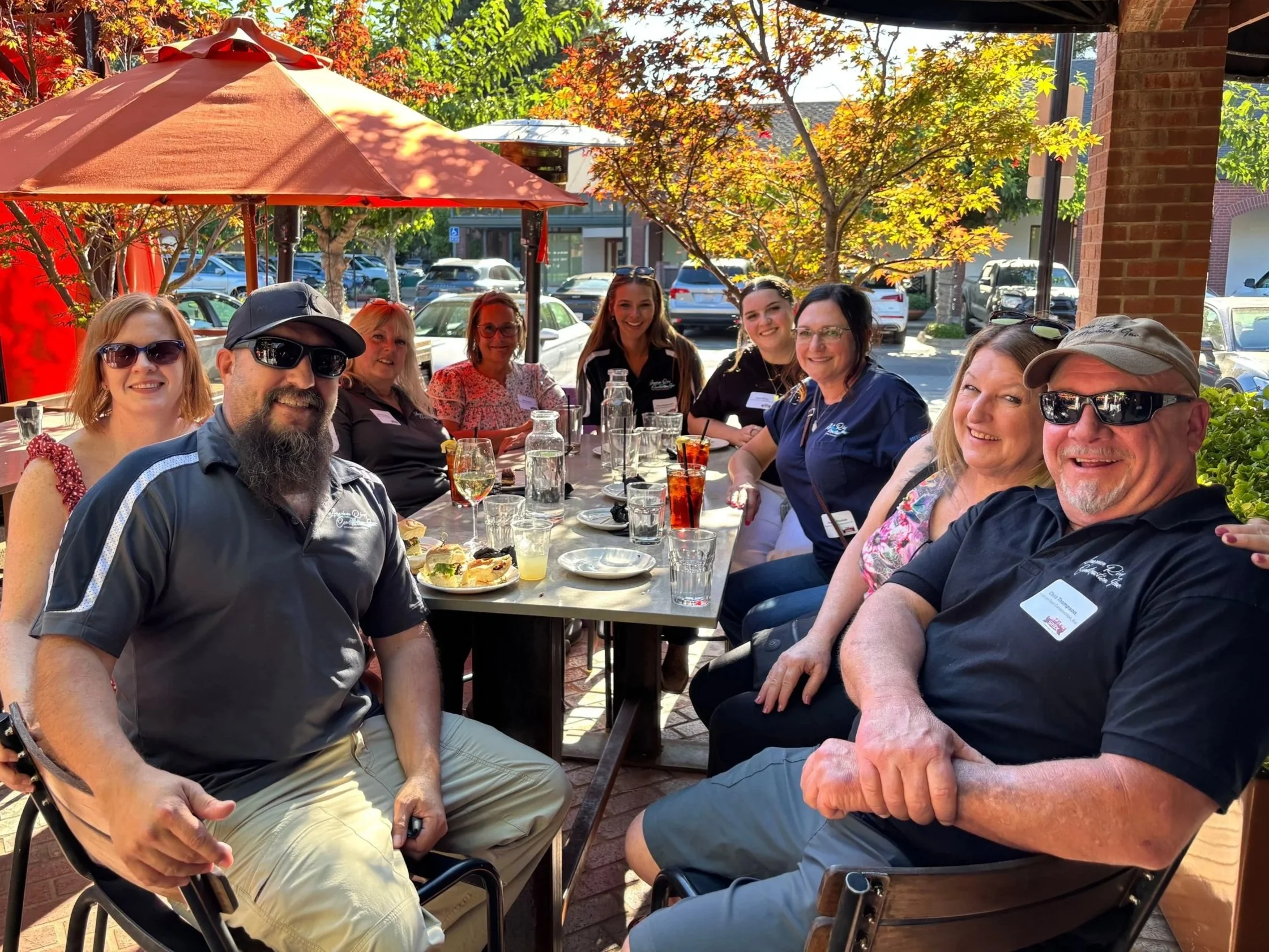 A group of people are sitting at a table outdoors during daytime, smiling, with glasses and plates of food in front of them. There are umbrellas and trees with autumn leaves in the background.