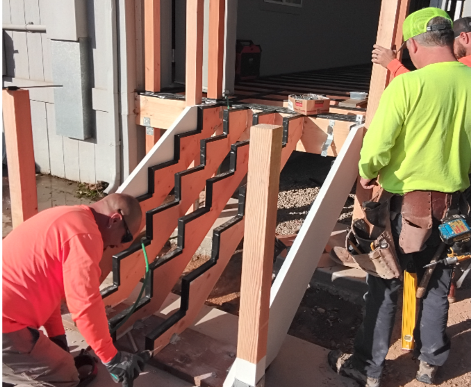Construction workers installing black zigzag stair stringers on a wooden staircase frame.