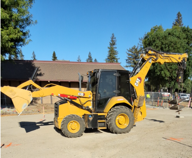 A yellow Caterpillar backhoe loader parked at a construction site on dirt ground with trees and a building in the background.