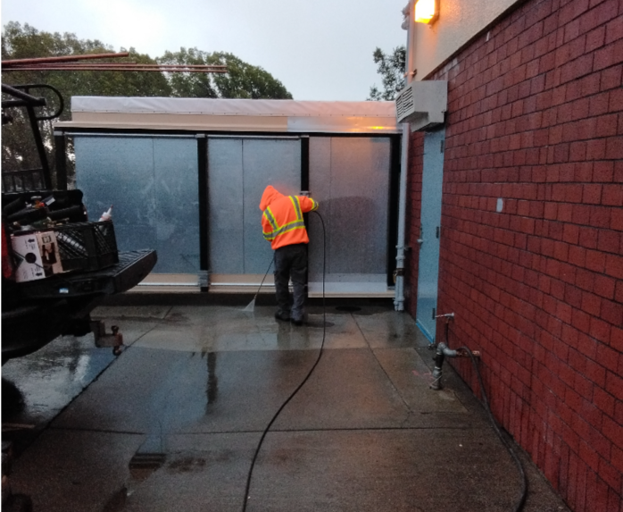 Worker in safety gear pressure washing a large metal door outdoors at dusk.
