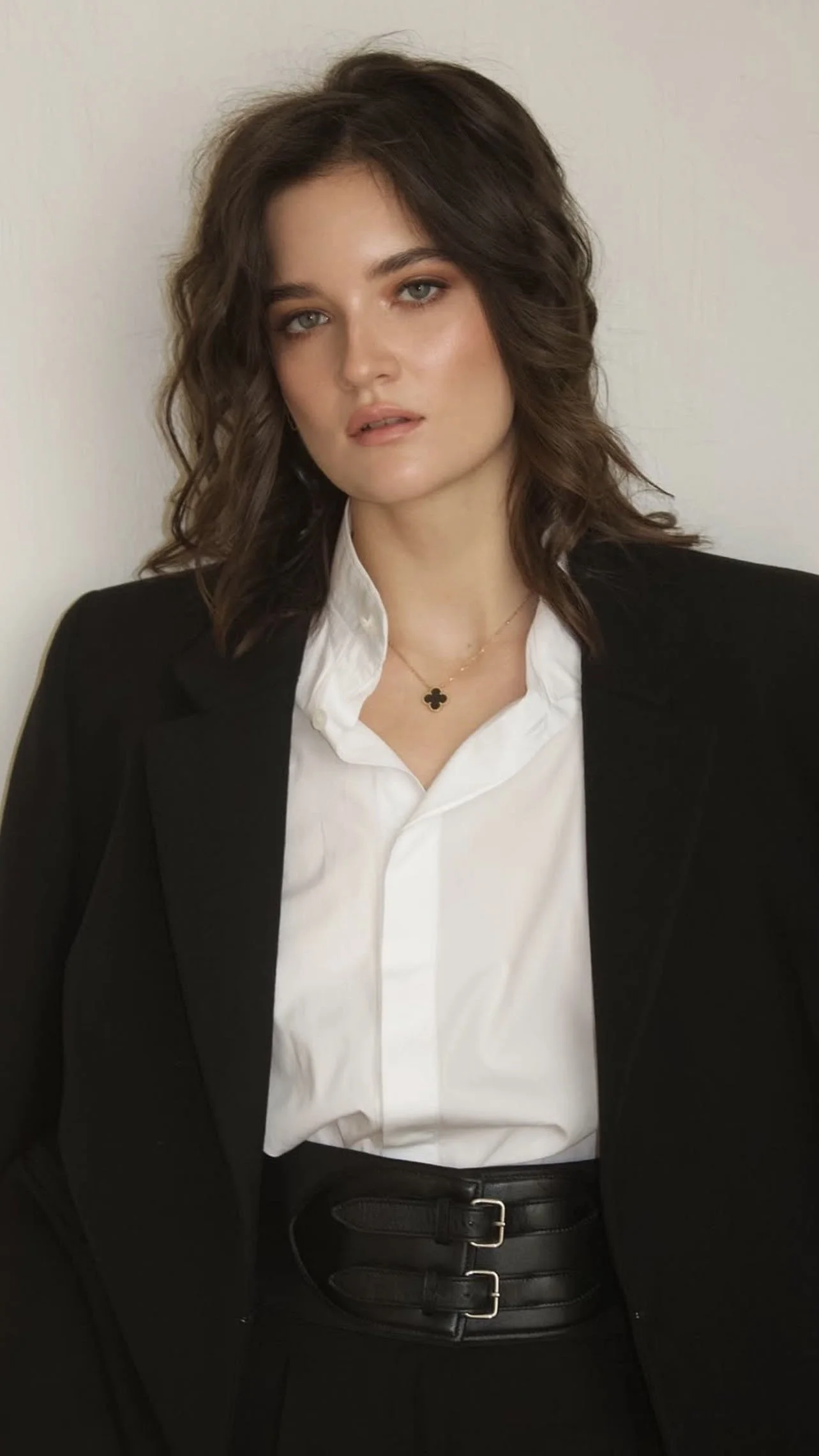A woman with wavy brown hair and blue eyes wearing a black blazer, white shirt, a gold necklace with a black flower pendant, and a wide black belt, standing against a plain background.