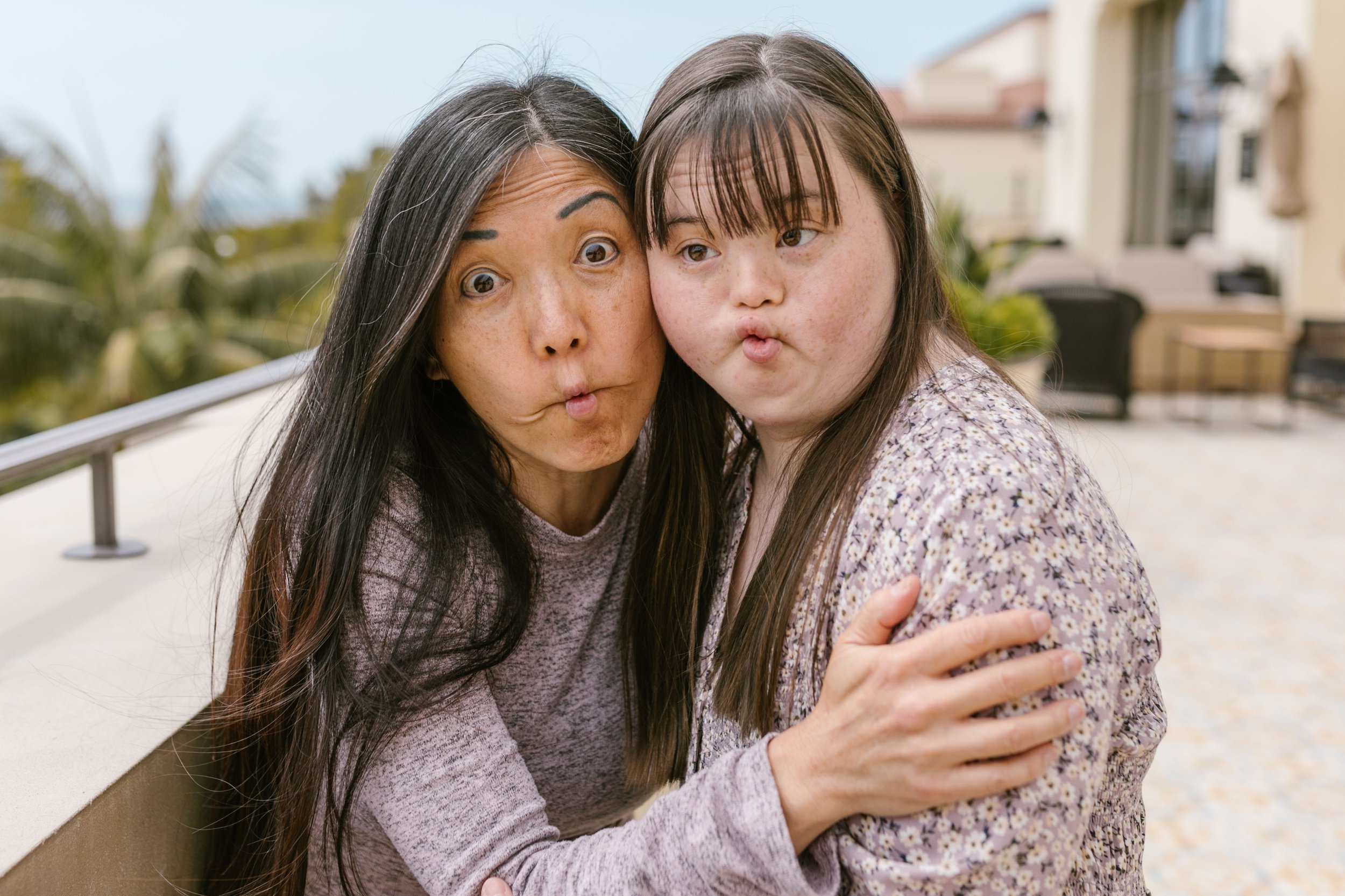 Two women, one with Asian features and long black hair, and the other with light skin and brown hair, are sharing a hug outdoors. They are both making playful, puckered expressions and look surprised or curious. The background shows a residential area with houses, patio furniture, and greenery.